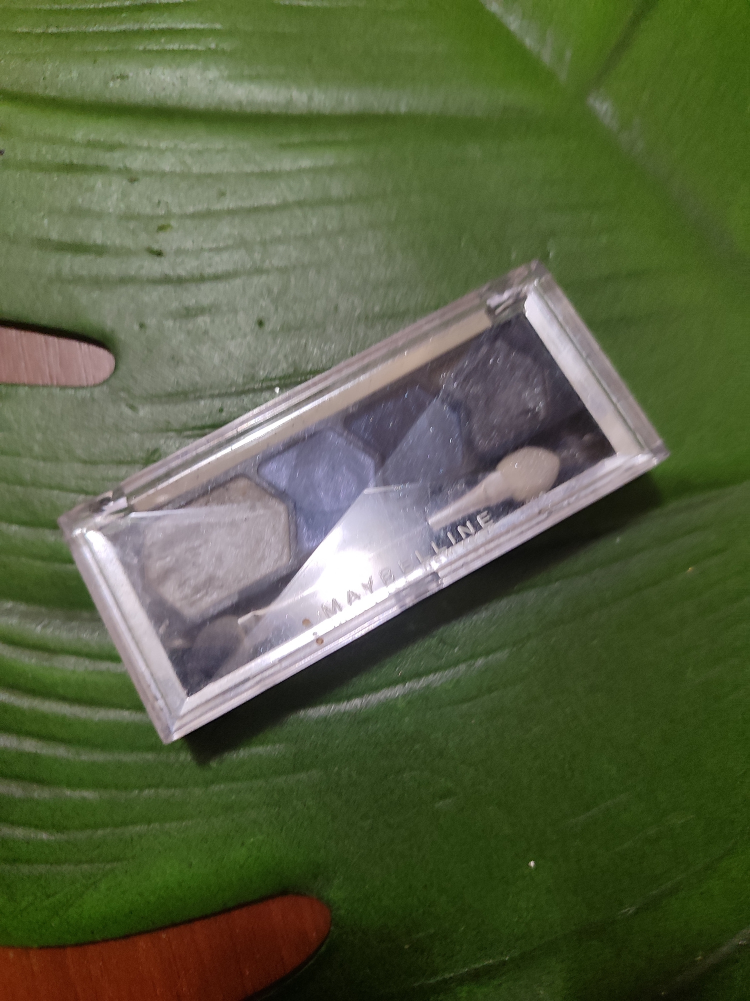 An open, rectangular, clear plastic eyeshadow palette containing four shades of gray and silver eyeshadow is positioned diagonally on a large, dark green, veined leaf, likely a Monstera. The palette is partially transparent, allowing some of the leaf's texture to be seen through it. The brand name "MAYBELLINE" is visible printed in white on the inside lid of the palette. A small, white applicator with a rounded tip rests inside the palette. The background is dominated by the textured surface of the leaf, with subtle light reflections suggesting an indoor setting. There are no people or signs of human activity in the immediate vicinity of the objects. The image appears to be a still life, focusing solely on the cosmetic item and its immediate surroundings.