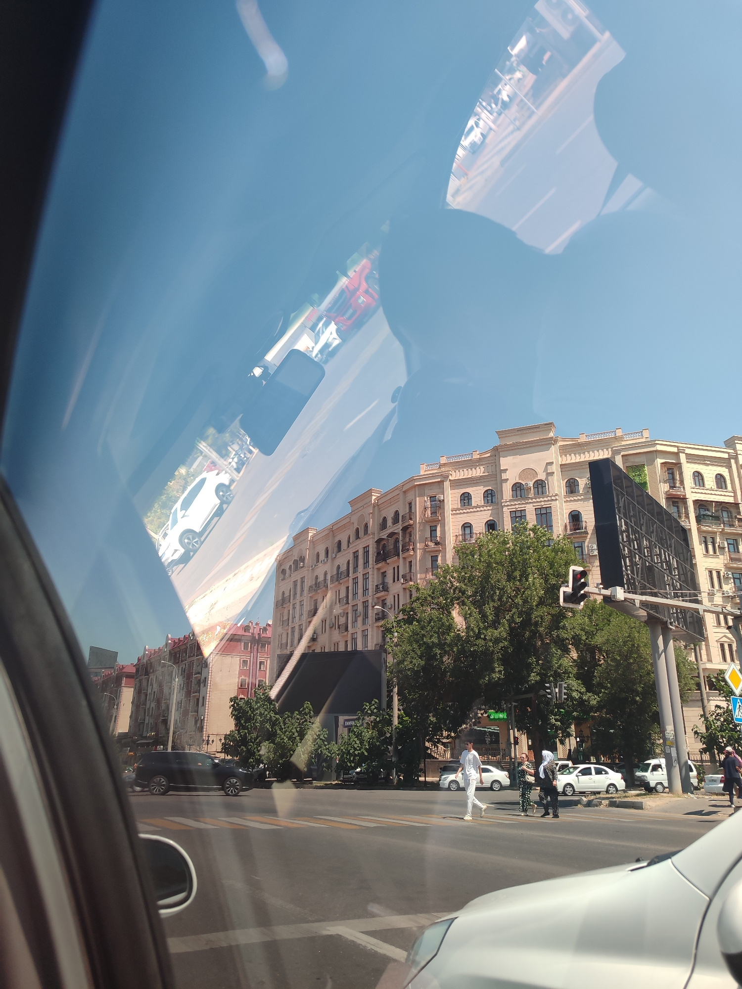 The image shows a view from inside a vehicle, looking out through the windshield. The scene is a city street in Tashkent, Uzbekistan. The time of day appears to be daytime, with bright sunlight.
In the foreground, the reflection of the vehicle's interior is visible on the windshield, including what appears to be a side mirror and part of the dashboard. Beyond the windshield, the street is lined with buildings. On the left side, there are multi-story buildings with red-tiled roofs. On the right side, there is a large, modern, light-colored building with arched windows and balconies, and a large black billboard.
Traffic is present on the street. A black SUV is driving on the road, and several other cars are visible, including white sedans and a white van. Pedestrians are crossing the street at a crosswalk. Two women in traditional Islamic head coverings are walking across the street, and another person in white is walking behind them. A traffic light is visible, showing a red signal. Trees are planted along the street, providing shade.
The sky is clear and blue. The overall impression is a bustling urban environment in Tashkent on a sunny day.
