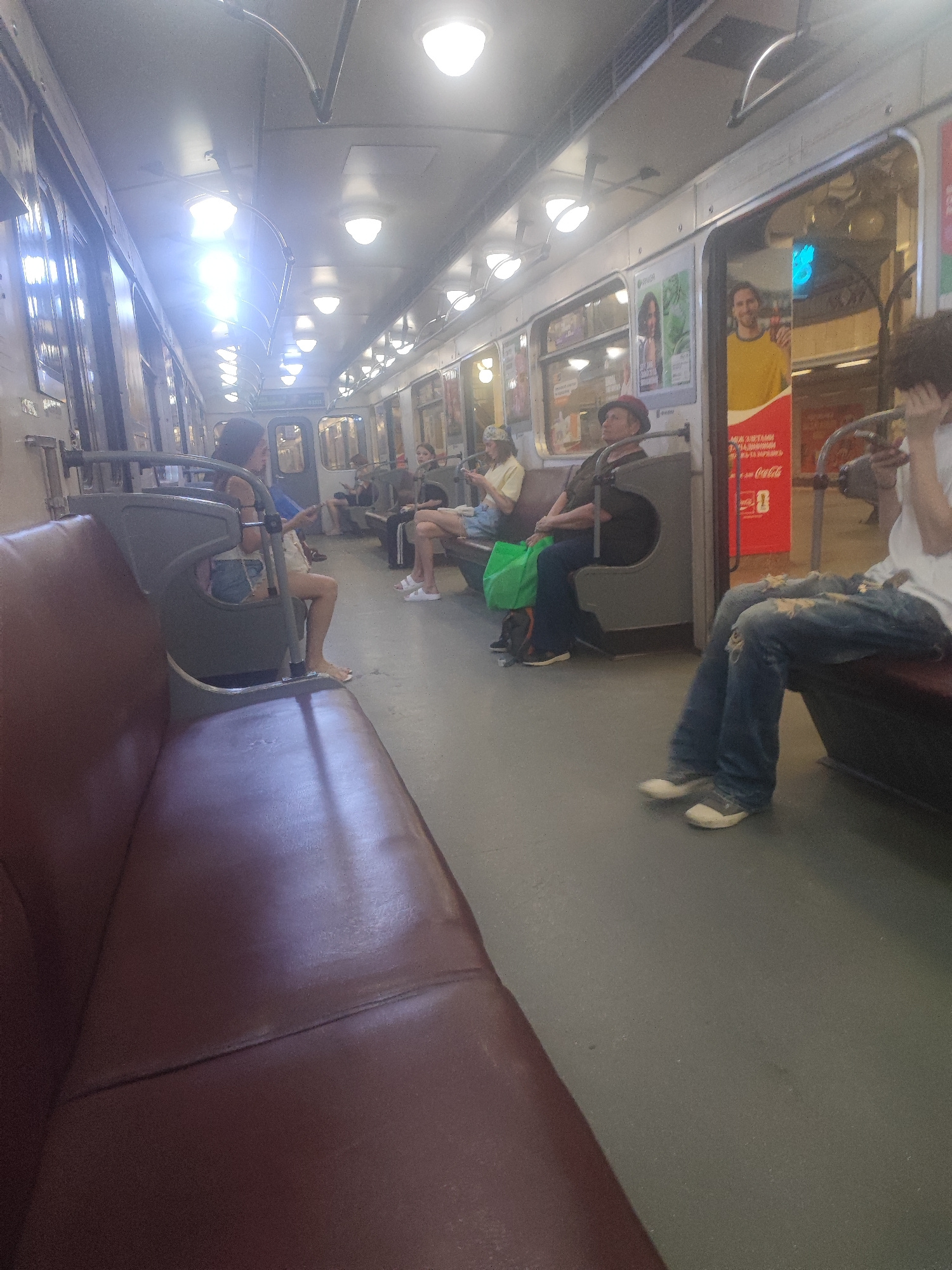 The image depicts the interior of a train car, likely a metro or subway, with several passengers. The setting is an indoor transit environment, illuminated by overhead fluorescent lights. The time of day is indeterminate, but the artificial lighting suggests it could be anytime, though it appears to be daytime given the bright exterior light filtering in from the windows.
The subjects are primarily people seated on the train. In the foreground, a portion of a maroon-colored seat is visible, suggesting it's a seating area. Further down the aisle, several individuals are seated. On the left, a woman with dark hair is seen wearing ripped denim shorts and a blue top, with her legs crossed and feet bare. To her right, a woman in a hat and dark clothing is looking upwards. Further down the car, a man in a dark shirt and hat is seated, holding a green bag. Another person, partially visible, is seated with their legs extended. On the far right, a person in ripped blue jeans and a white shirt is seated, with their hands resting on their lap.
The train car appears to be in motion or at a stop, with passengers going about their journeys. The mood seems to be mundane, typical of daily commuting. Notable details include the worn interior of the train car, with advertisements visible on the walls. One advertisement features a smiling man in a yellow shirt and text that appears to be in Cyrillic script. Below it, the Coca-Cola logo is visible. The floor of the train car is a neutral gray. The overall impression is of a crowded but ordinary public transportation scene in an urban setting. The location cue of "Kyiv, Ukraine" indicates this is a scene from that specific city.