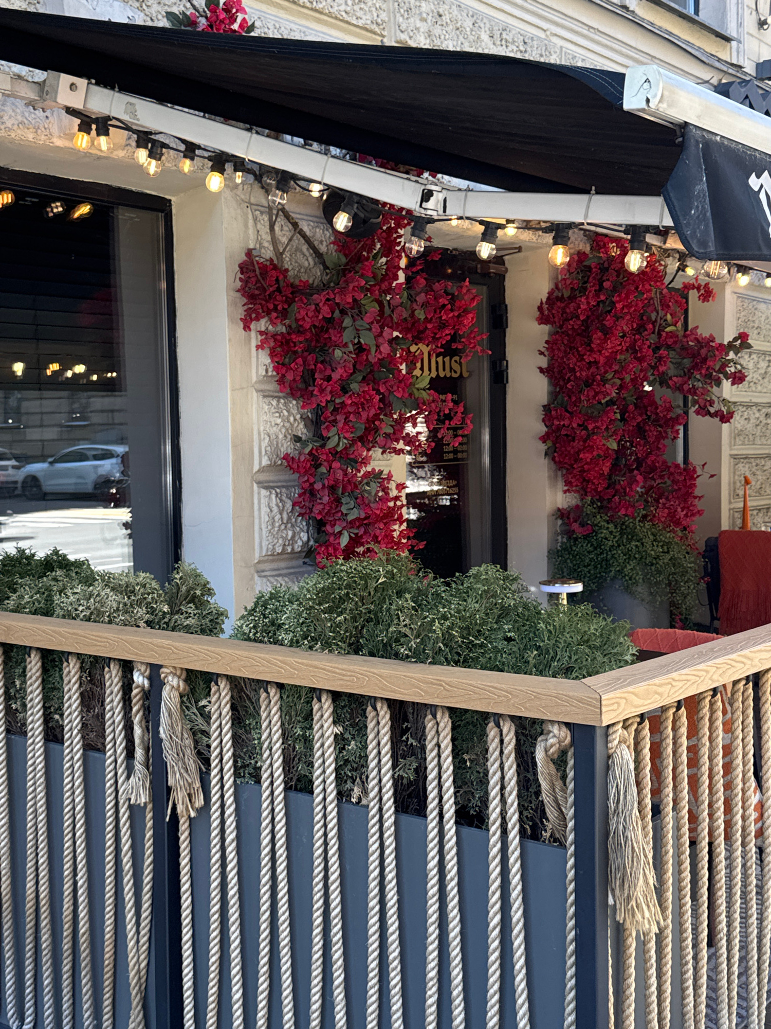 The image shows a close-up view of an awning over a storefront, adorned with strings of lights and artificial red flowers. The awning is dark blue and made of fabric, with a metal frame. Below the awning, a window reveals the interior of the establishment, with some of the lights reflecting in the glass. The storefront appears to be decorated with more red flowers and greenery, suggesting an outdoor or semi-outdoor seating area. To the right of the storefront, a decorative wooden structure with thick ropes stretched across it is visible. The ropes are beige and appear to be made of natural fibers, creating a textured, nautical-inspired element. The lighting in the image suggests it might be evening or a dimly lit day, as the string lights are illuminated. The overall scene evokes a cozy and inviting atmosphere, perhaps a cafe, restaurant, or shop.
There is visible text on the awning, though it is partially obscured and difficult to read clearly. It appears to be a logo or name, possibly in white lettering. The text "Just" is also visible in the reflection of the window.