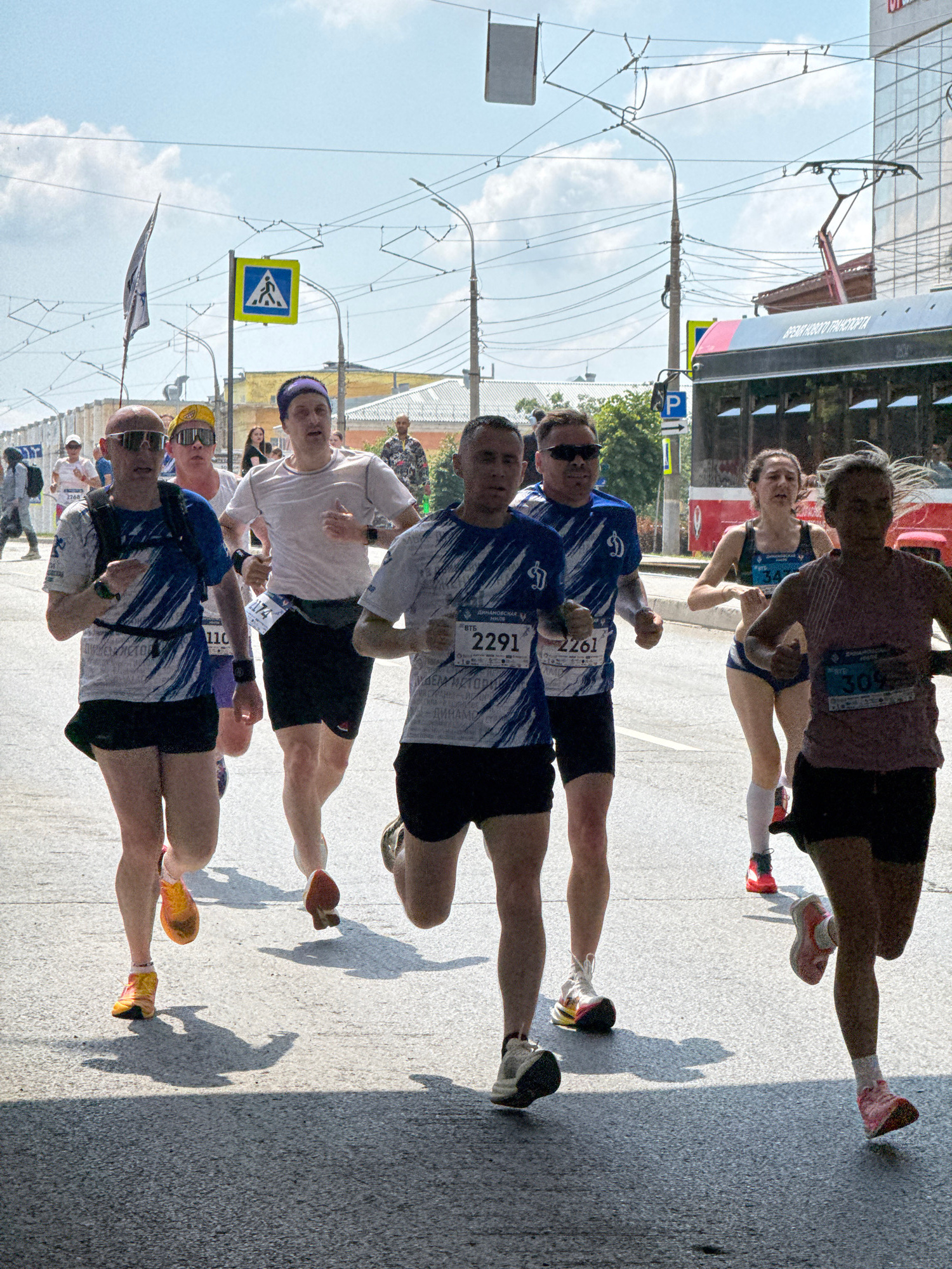 The image captures a marathon race taking place on a sunny day in the city of Izhevsk, Russia. Several runners are depicted in mid-stride, showcasing various athletic attire and race bibs. The foreground is dominated by the legs and torsos of the runners, with some displaying determined facial expressions. In the background, a modern red bus with text indicating "ВРЕМЯ НОВОГО ТРАНСПОРТА" (Time for new transport) is visible, along with power lines, streetlights, and buildings that suggest an urban environment. A pedestrian crossing sign is also present, reinforcing the city setting. The ground is paved with asphalt, and shadows cast by the runners and other elements indicate the bright daylight. The overall scene conveys the dynamic energy of a sporting event in a public urban space.