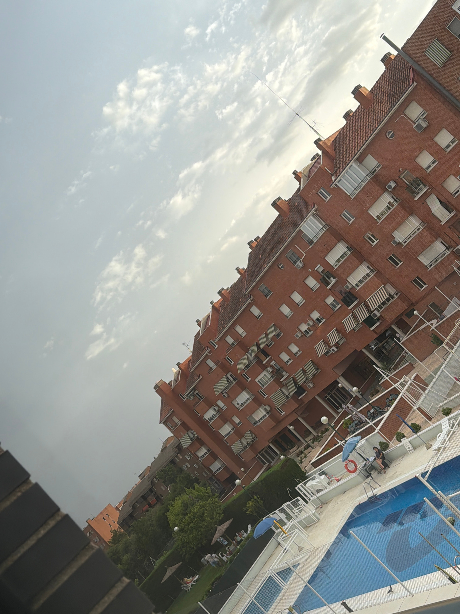 An angled, eye-level medium shot captures a brick apartment building overlooking a swimming pool and surrounding green space. The building, with its red tiled roof and numerous windows, dominates the right side of the frame. Several stories are visible, many with balconies featuring awnings. A thin antenna extends vertically from the rooftop.
Below the building, a blue swimming pool with a white border and fence is visible. The water reflects the sky, showing a faint shadow pattern. A few lounge chairs and umbrellas are scattered on the sandy area surrounding the pool. Two people are near the pool; one is seated on a chair, and another is standing.
In the background, more buildings and trees can be seen. The sky above is a mix of light gray clouds and patches of lighter sky, suggesting an overcast or partly cloudy day. The overall impression is of a residential area with leisure facilities.
