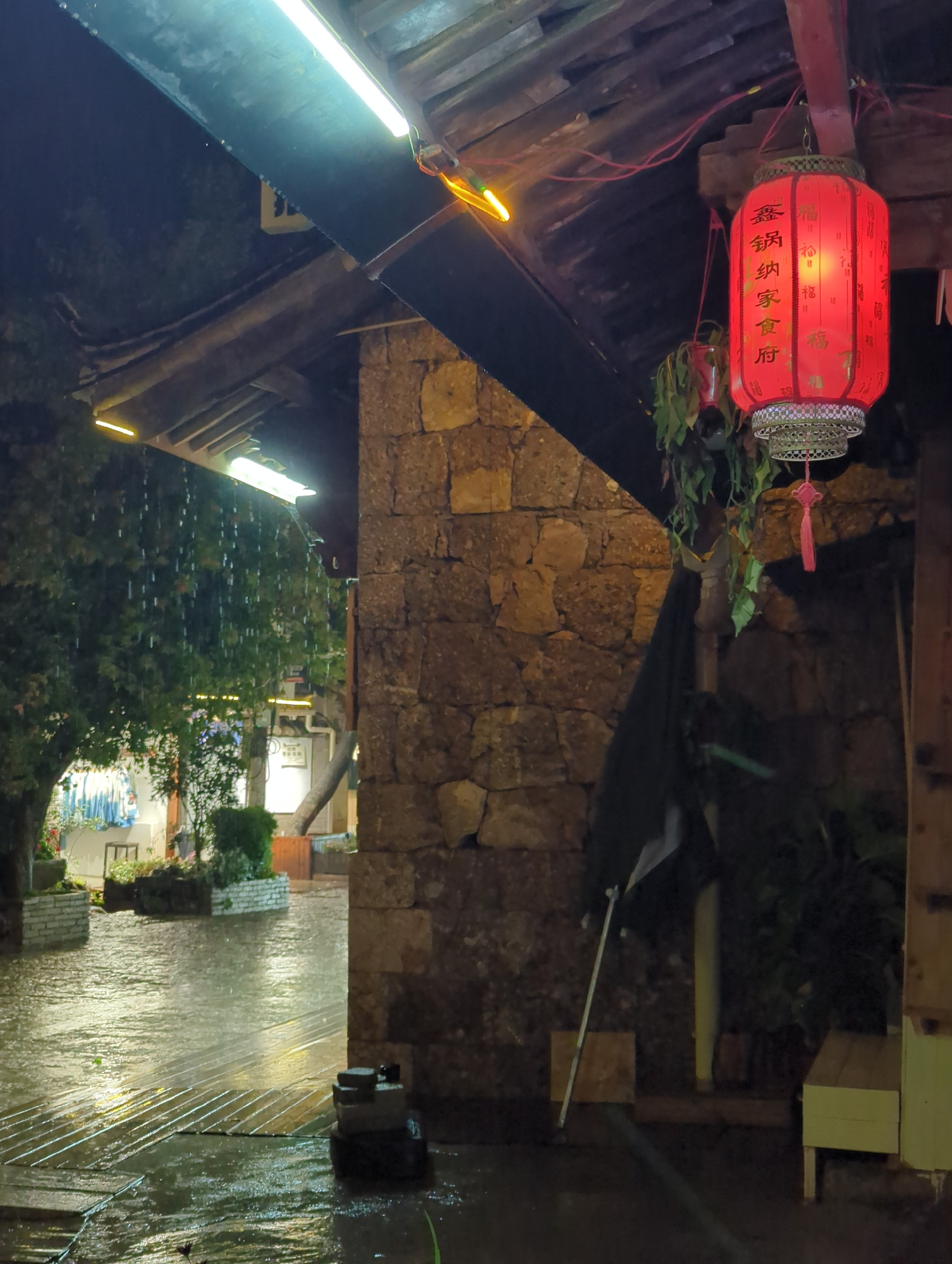 The image depicts an outdoor scene at night in Yulong Nakhi Autonomous County, China, during a rainfall. The foreground shows a wet, paved area with wooden planks, reflecting the ambient light. To the right, a stone wall forms part of a building structure, with a wooden bench visible. Above, a wooden overhang supports a red lantern with Chinese characters, emitting a warm glow. A black umbrella leans against the wall, and hanging plants add greenery. To the left, trees with water droplets falling from their leaves are visible, along with shopfronts displaying clothing. The overall atmosphere is wet and dimly lit, characteristic of a rainy night in a town.