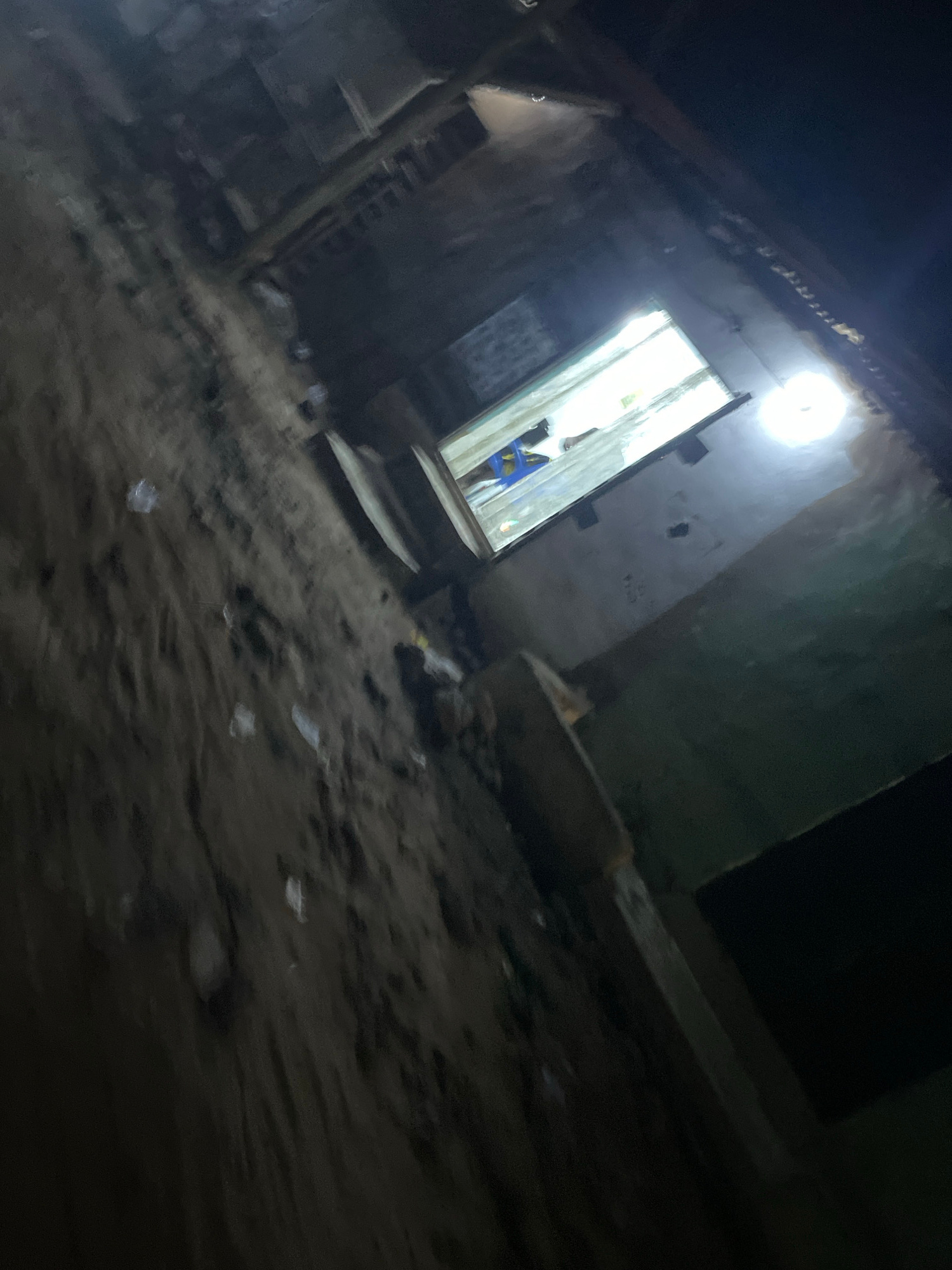 This image captures an interior view of a dilapidated, multi-story structure in Lokoja, Nigeria. The scene appears to be an abandoned or neglected building, possibly a former residential or commercial property, with visible signs of decay and disrepair. The perspective suggests the photographer is looking down from an upper level, observing the area below through an opening or a window.
The lower portion of the frame is dominated by rough, textured surfaces that resemble packed earth or concrete, with debris scattered across it. This debris includes what appear to be plastic wrappers and other refuse, indicating a lack of maintenance. Above this, a prominent opening or window reveals another section of the interior. This opening is framed by what looks like exposed wooden beams and rudimentary construction materials.
Within this windowed area, a distinct human presence is visible. A person, or possibly two, are seen interacting with objects. One individual appears to be wearing a blue and yellow garment. They are positioned near a bright light source emanating from outside the frame, likely a window or an opening that allows significant daylight to penetrate the dim interior. The light creates a strong contrast, illuminating this section while leaving much of the surrounding structure in shadow.
The overall environment is dark and grimy, with a strong sense of abandonment. The architectural style suggests a building that is not modern, possibly an older construction that has fallen into disuse. The lighting conditions, with the bright light from the opening and the general dimness elsewhere, contribute to a somewhat eerie or unsettling atmosphere. The orientation of the image, tilted from the horizontal, adds to the dynamic and perhaps disorienting feel of the scene.