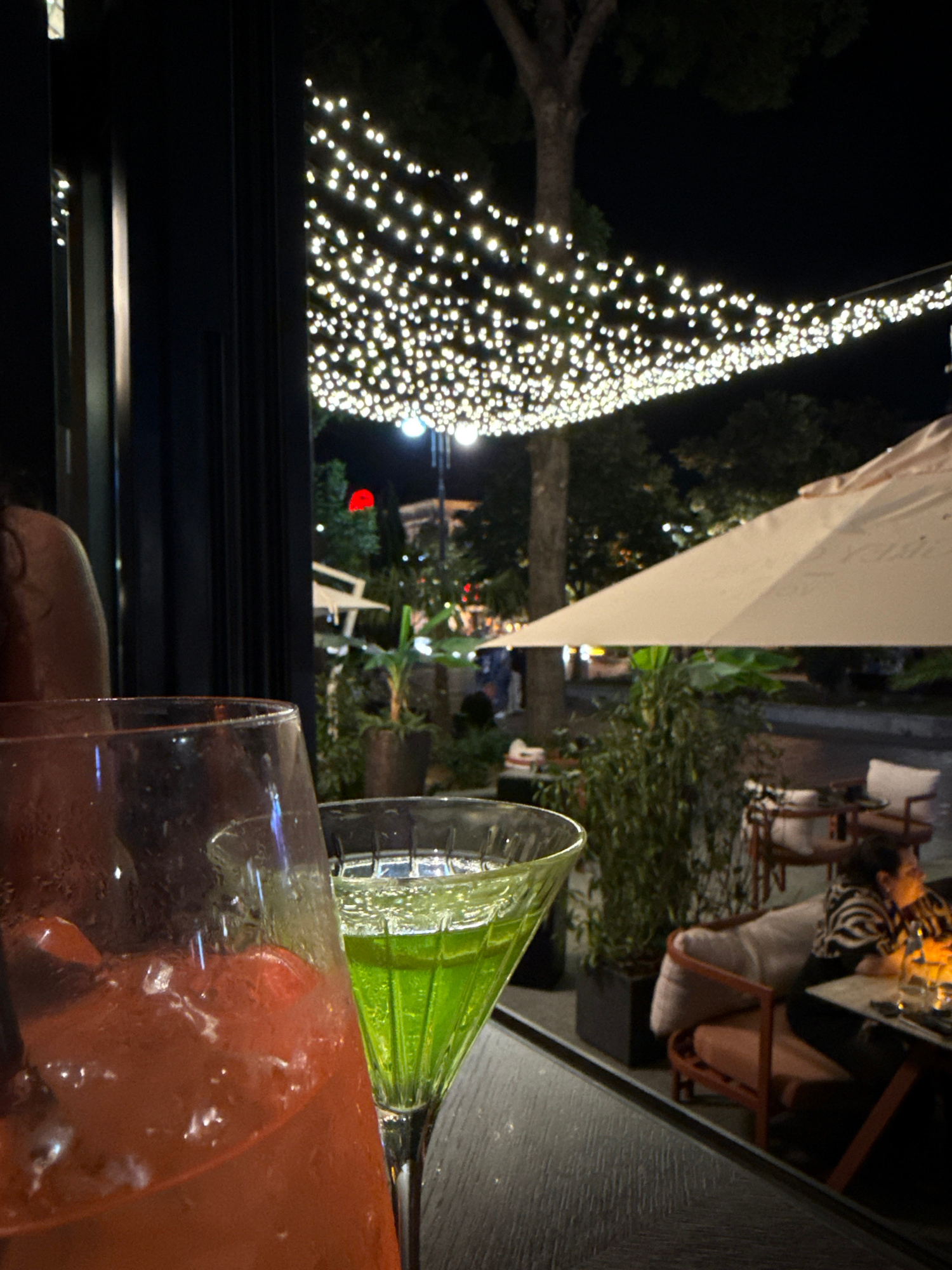 In an outdoor evening setting, a close-up view shows two drinks on a dark, textured surface. The drink on the left is in a large pitcher, filled with a red liquid and ice. To its right, a martini glass holds a vibrant green liquid. In the background, a string of white lights hangs across the scene, illuminating trees and the night sky. Further back, a large, light-colored umbrella shades outdoor seating where a person wearing a black and white striped top is visible, interacting with someone seated at a table. The environment suggests a lively urban nightlife scene with ambient lighting and other patrons.