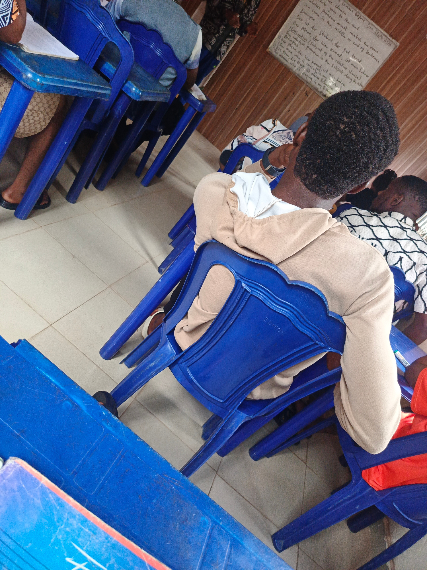 This media file captures an indoor scene, likely a classroom or meeting hall, with several people seated on blue plastic chairs. The focus is on the back of a young person wearing a tan hoodie and light-colored pants. They are seated at a blue plastic desk, with a notebook open in front of them. Another person, wearing a black and white patterned shirt, is visible in the background, also seated.
The scene appears to be a learning environment, as evidenced by a whiteboard with handwritten text in German visible on the wall. The text discusses grammar, specifically relative clauses. The handwriting is clear, and the whiteboard serves as a notable detail, suggesting an educational activity is taking place. The overall lighting is somewhat dim, hinting that it might be daytime but without direct sunlight. The tiled floor and the wooden paneled wall provide further clues to the setting.
There are no discernible emotions displayed by the subjects, as most of them are only partially visible or their faces are not the focus. The presence of notebooks and the whiteboard strongly indicates a class or study session. The blue plastic chairs and desks suggest a functional rather than a decorative setting, common in educational institutions or community centers. The time of day and weather are not evident from the image.