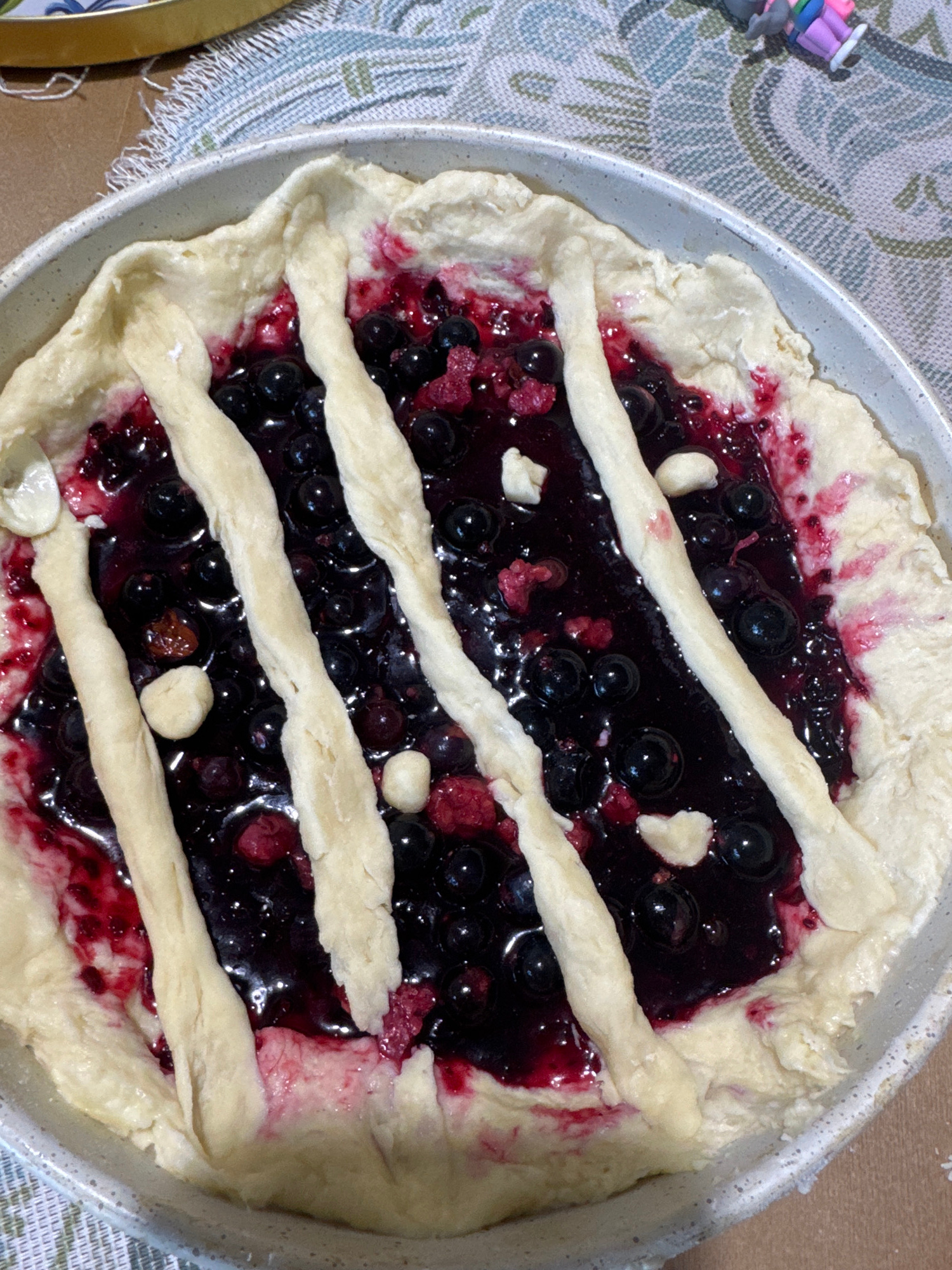 An overhead, close-up shot shows a rustic berry galette in a round, light-gray baking dish, set on a textured tablecloth with a green, white, and blue geometric pattern. The galette's crust is unbaked and folded over the edge of the pie dish, with a lattice of pastry strips covering the dark berry filling, which appears to be composed of blueberries and raspberries. Some of the berries have burst, releasing their juices, and a few small, light-colored pieces, possibly chunks of dough or cheese, are scattered on top. The background of the image shows a corner of a golden tray and a small, colorful toy figure on the tablecloth. The scene appears to be indoors, likely in a kitchen or dining area, but no specific location in Kokshetau, Kazakhstan, can be identified from the visual information provided.