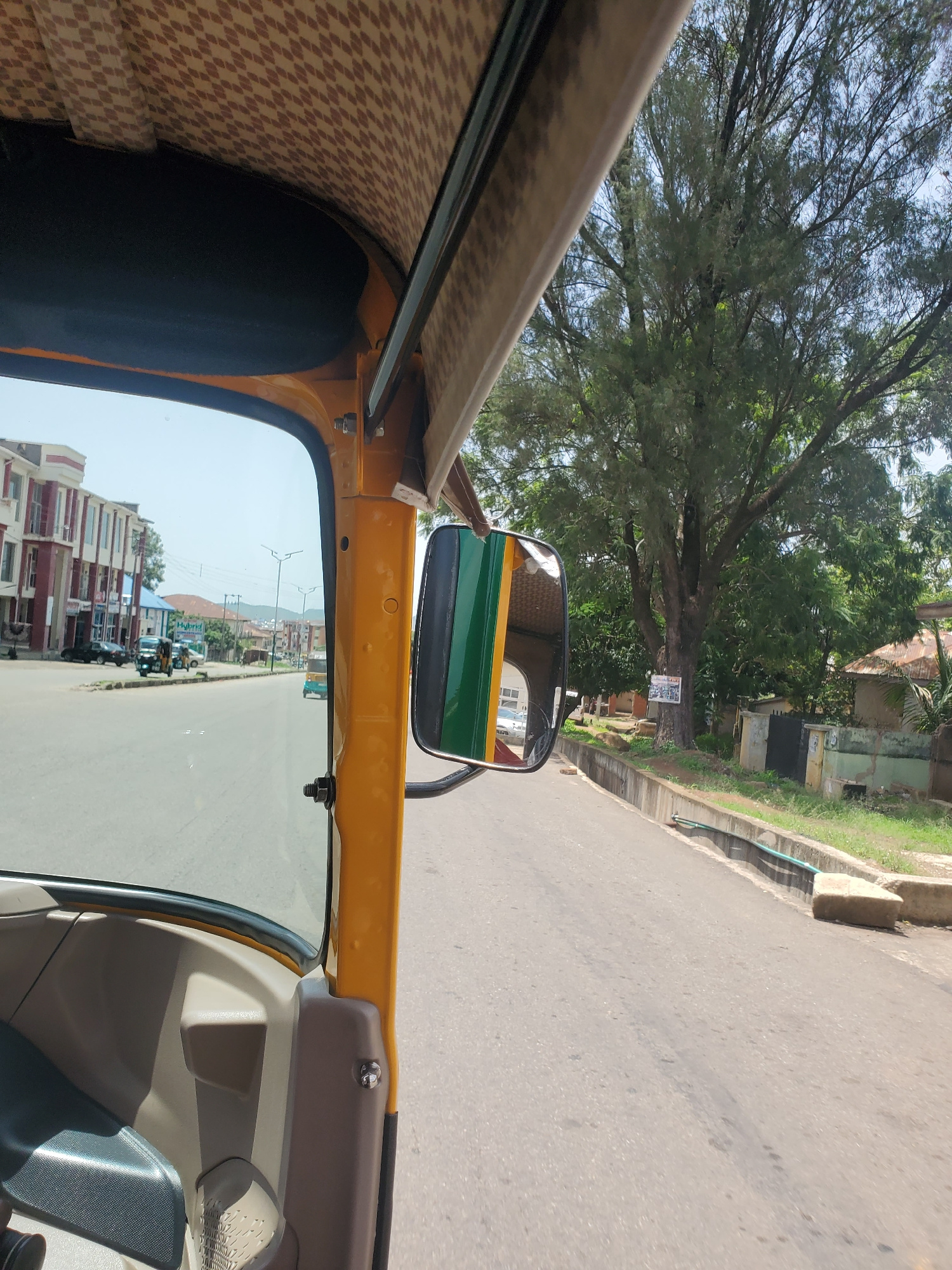 The image captures a view from inside a yellow auto-rickshaw, looking forward and slightly to the right. The interior of the auto-rickshaw features brown and beige patterned upholstery. The exterior side mirror, reflecting the green and yellow colors of the auto-rickshaw, is visible.
The scene outside shows a road with a sidewalk bordered by grass and a drain. Tall trees with dense foliage line the road, casting shadows. In the distance, buildings with red and white facades are visible, along with utility poles and wires. Several other vehicles, including motorcycles and what appear to be more auto-rickshaws, are on the road. The sky is bright, suggesting it is daytime, likely mid-morning or afternoon. The weather appears clear and sunny.
A white sign with some text is attached to a tree, but the text is not legible. The overall impression is of a street scene in a tropical or subtropical urban environment, possibly in Jos, Nigeria, given the context. The image is taken from a moving vehicle, indicated by the slight blur of the background and the perspective. No people are clearly discernible as subjects, but their presence is implied by the vehicles and the urban setting.