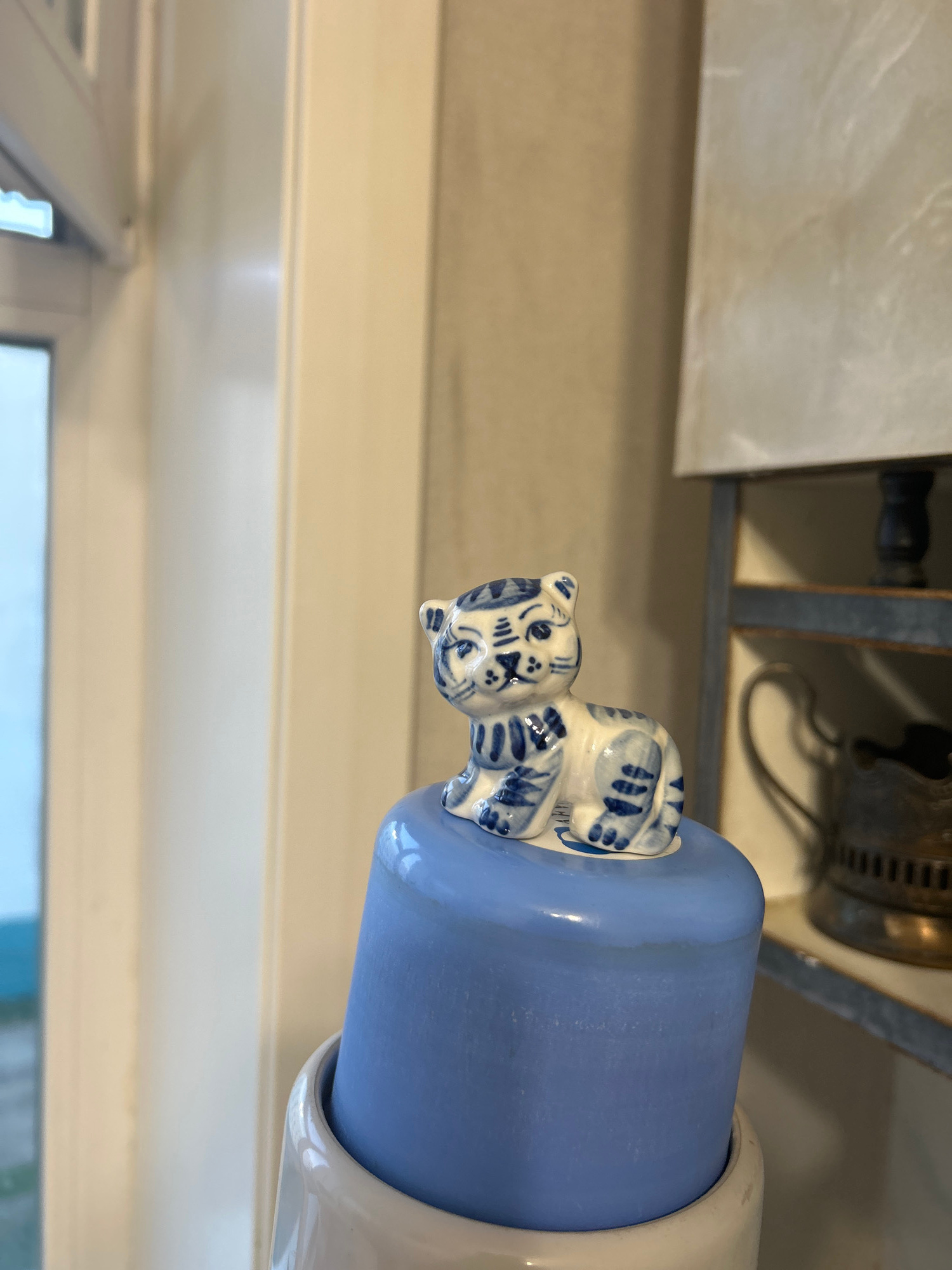 The image shows a close-up of a ceramic figurine of a cat with blue and white markings, resting on a blue cylindrical object. The cat is depicted in a seated position, with its paws tucked in. The figurine appears to be a decorative item. The background is blurred, but suggests an indoor setting with some shelving or cabinetry visible. The lighting appears to be artificial, casting soft shadows. There is no visible text in the image. The image is oriented vertically.