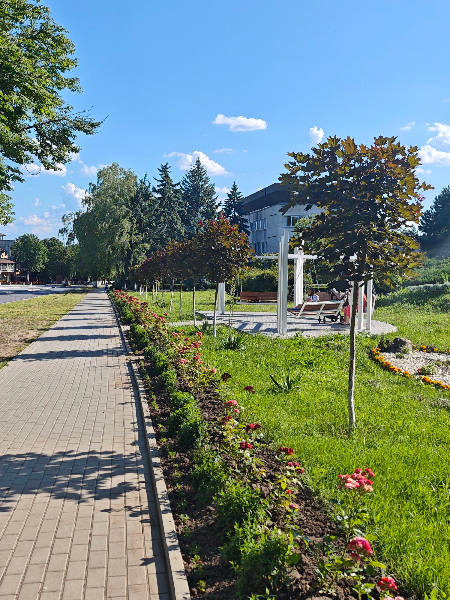A sunlit, wide paved walkway with gray rectangular bricks extends diagonally from the lower left to the mid-right of the image. The walkway is bordered on the right by a row of meticulously maintained flower beds filled with blooming red roses and green foliage. Beyond the flower beds, a vibrant green lawn stretches towards the background. In the middle ground, a modern white gazebo-like structure houses two swings. On one swing, a young girl with blonde hair and a light-colored top is seated, facing left. Next to her, another person is partially visible, also seated. A stroller is placed in front of the swing. To the left of the walkway, a street is visible with parked cars and buildings, including a restaurant with outdoor seating. The background features a line of tall, mature trees, including evergreen and deciduous varieties, and a modern, light-colored building with multiple windows. The sky is a clear, bright blue with a few scattered white clouds. The scene depicts a well-kept urban park area on a sunny day.