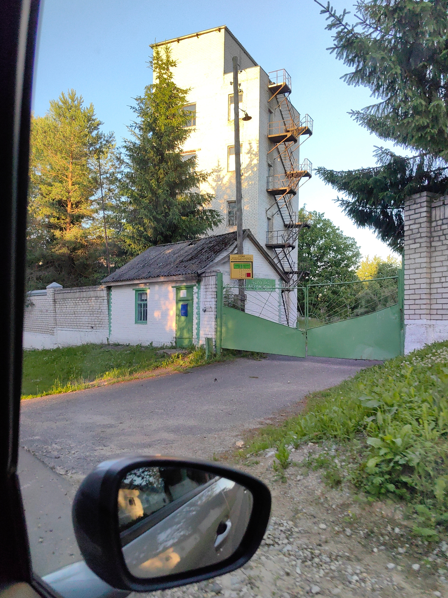 The image shows a view from inside a car, looking out towards a building complex. The scene is set during the late afternoon or early evening, as indicated by the long shadows and warm lighting. The main building is a tall, white, multi-story structure with a rusted metal fire escape attached to its side. In front of this building is a smaller, white structure with a green door and window, and a weathered roof. A green metal gate, partially open, is visible in the foreground, leading into the complex.
The environment consists of a paved road, a grassy area with some weeds, and a white brick fence. There are several mature evergreen trees scattered around the property. The sky is clear and blue. A side-view mirror of the car is visible in the lower left corner of the image, reflecting a blurred image of what appears to be a dog or animal inside the car.
The location is identified as Дзяржынскі сельскі Савет, Belarus. The overall impression is that of an industrial or institutional site, possibly an older facility, given the architecture and the weathered appearance of some elements. There are no people visible in the scene.