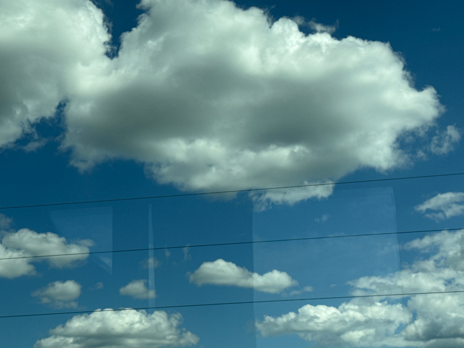 The image shows a bright blue sky filled with white, fluffy clouds. Three thin, dark horizontal lines are visible, suggesting they are wires or reflections across the sky. The clouds vary in size and density, with some appearing as large, dense masses and others as smaller, more scattered formations. The lighting indicates it is daytime. There is no indication of the city of Jaunolaine, Latvia within the visual content of the image.