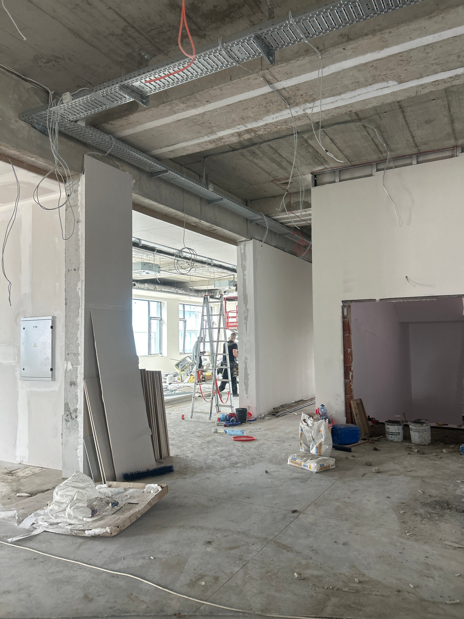 The image depicts an interior construction site, likely in Sofia, Bulgaria, showing ongoing renovation work. In the foreground, the concrete floor is partially covered with dust and debris. To the left, a white electrical panel is mounted on a concrete pillar. Several large, flat sheets of drywall are leaning against the pillar. Further into the scene, a doorway leads into a room with partially finished walls.
The ceiling features exposed concrete beams with extensive metal cable trays running along them, supporting numerous electrical wires. In the middle ground, a tall ladder is positioned, and two construction workers are visible. One worker, wearing a dark shirt and pants, is on the ladder, and another worker, in a light-colored shirt and dark pants, stands nearby.
Various construction materials and tools are scattered around the floor, including bags of cement, buckets, and a broom. The overall environment suggests an active construction or renovation project in progress. The lighting appears to be a mix of natural light from windows in the background and overhead artificial lighting.