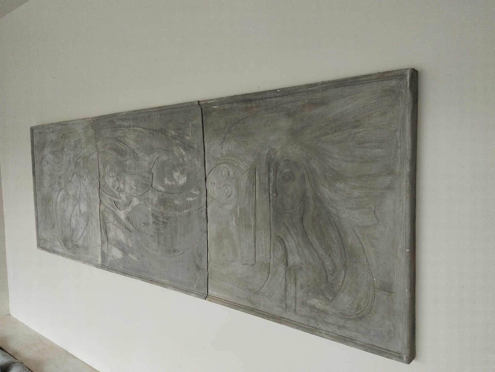 A diptych artwork, composed of two rectangular panels joined vertically, is displayed on a plain, light-colored wall. The artwork itself is textured and predominantly gray, with carved or raised relief elements depicting abstract forms. The left panel features swirling lines and shapes that suggest figures or facial features. The right panel also exhibits similar abstract, flowing designs, with distinct outlines of what could be interpreted as figures or faces. The artwork appears to be framed by a thin, darker border. The wall is a neutral, off-white color, with a subtle texture visible. The lighting is even, casting minimal shadows. The composition is centered on the artwork, with the wall extending to the edges of the frame. The environment appears to be an indoor setting, possibly a gallery or a home interior, with no other objects or furniture visible. The image provides no specific geographic markers to pinpoint a location within Kuta Selatan, Indonesia.