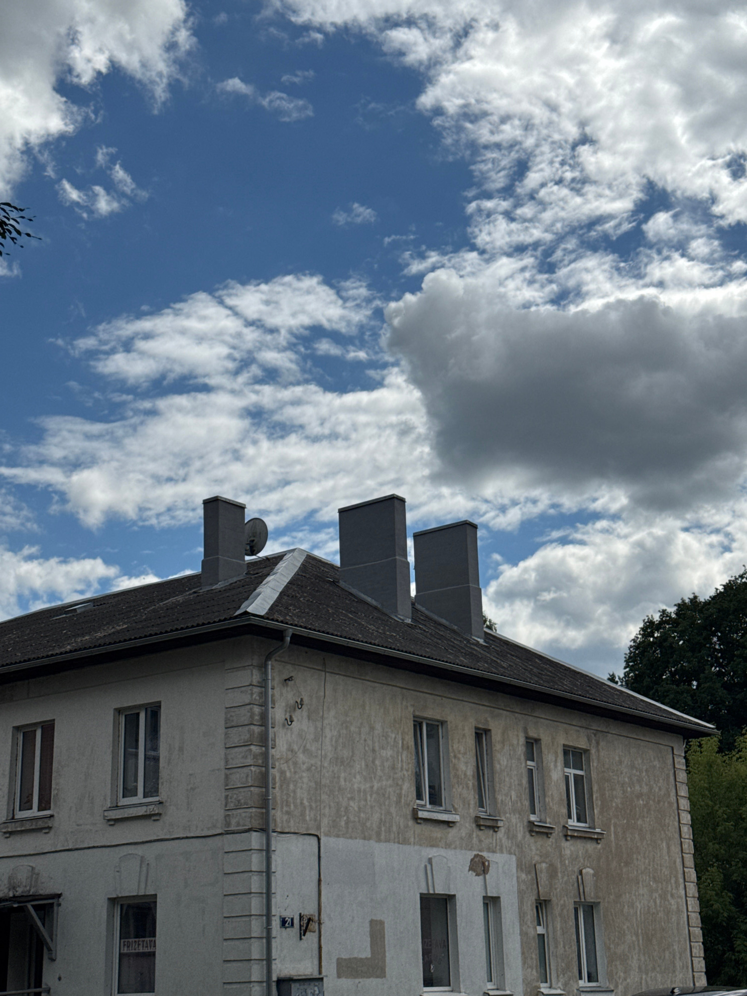 This image captures a close-up of a light-colored, weathered building with multiple windows and three prominent gray chimneys on its dark roof. The sky above is a vibrant blue, dotted with scattered white clouds of varying sizes and densities. In the lower left corner, part of a tree with dark leaves is visible. The building features distinct architectural details such as decorative stone pilasters on its corners and a gutter system along the roofline. A satellite dish is mounted on the roof near one of the chimneys. The windows are framed in white, and some appear to be closed. On the side of the building, a sign with the number "21" is visible, along with some electrical conduits and fixtures. A window on the lower floor has a sign that reads "FRIZETAVA". The overall impression is of an older building, possibly residential or commercial, set against a partly cloudy sky. This building is located at Rīgas iela 21 in Olaine, Latvia.