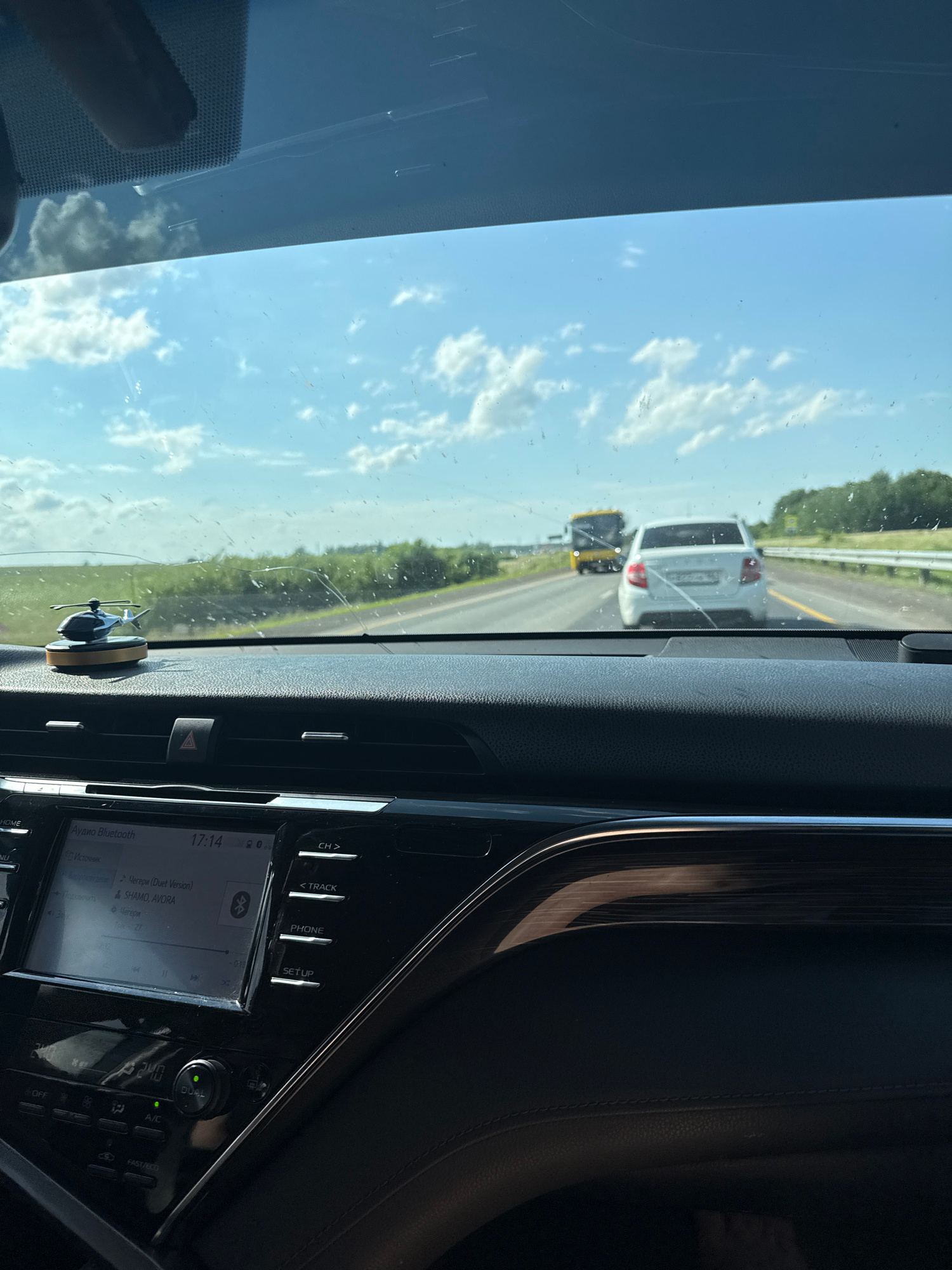 This media file captures a view from inside a car, looking out through the windshield. The scene is a highway with two other vehicles ahead. The car in front is a white SUV, followed by a yellow truck. The sky is clear and blue with scattered white clouds, suggesting a sunny day. The interior of the car is partially visible, showing part of the dashboard with a touchscreen display and various buttons. The display shows the time as 17:14 and indicates an "Audio Bluetooth" connection with song titles and artist names in Russian. A small decorative helicopter is mounted on the dashboard. The overall setting is a road trip on a highway under a bright sky.