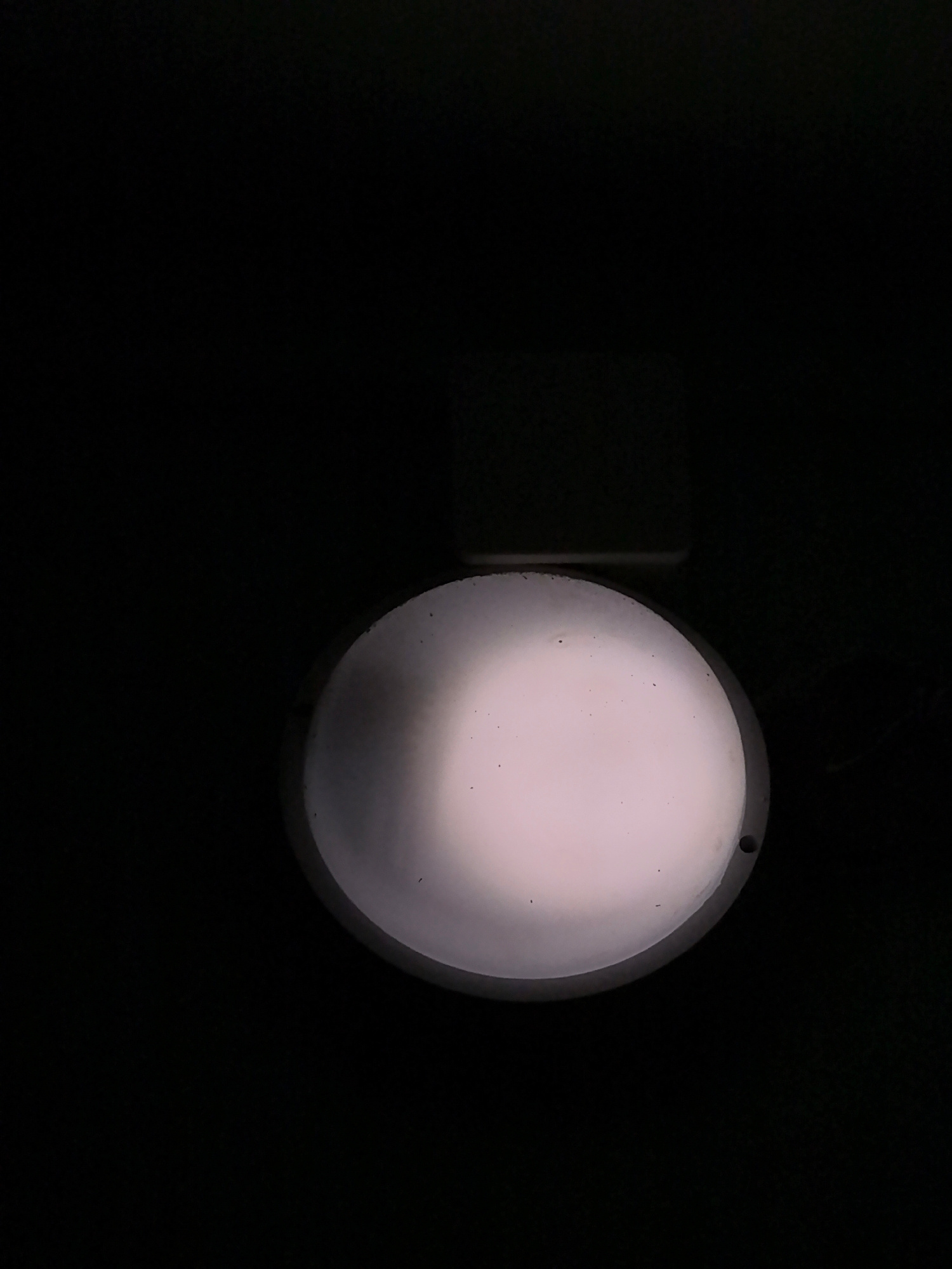 A circular, white, translucent object is illuminated from behind, creating a soft glow. The object is dark on the left side, gradually becoming lighter towards the right. The background is entirely black, suggesting a dark or nighttime environment. There appear to be small dark specks on the surface of the illuminated object. The object is viewed from a slightly angled, high perspective. A thin, rectangular, dark object is visible above the circular object. A small, dark circular indentation is present on the right edge of the circular object.