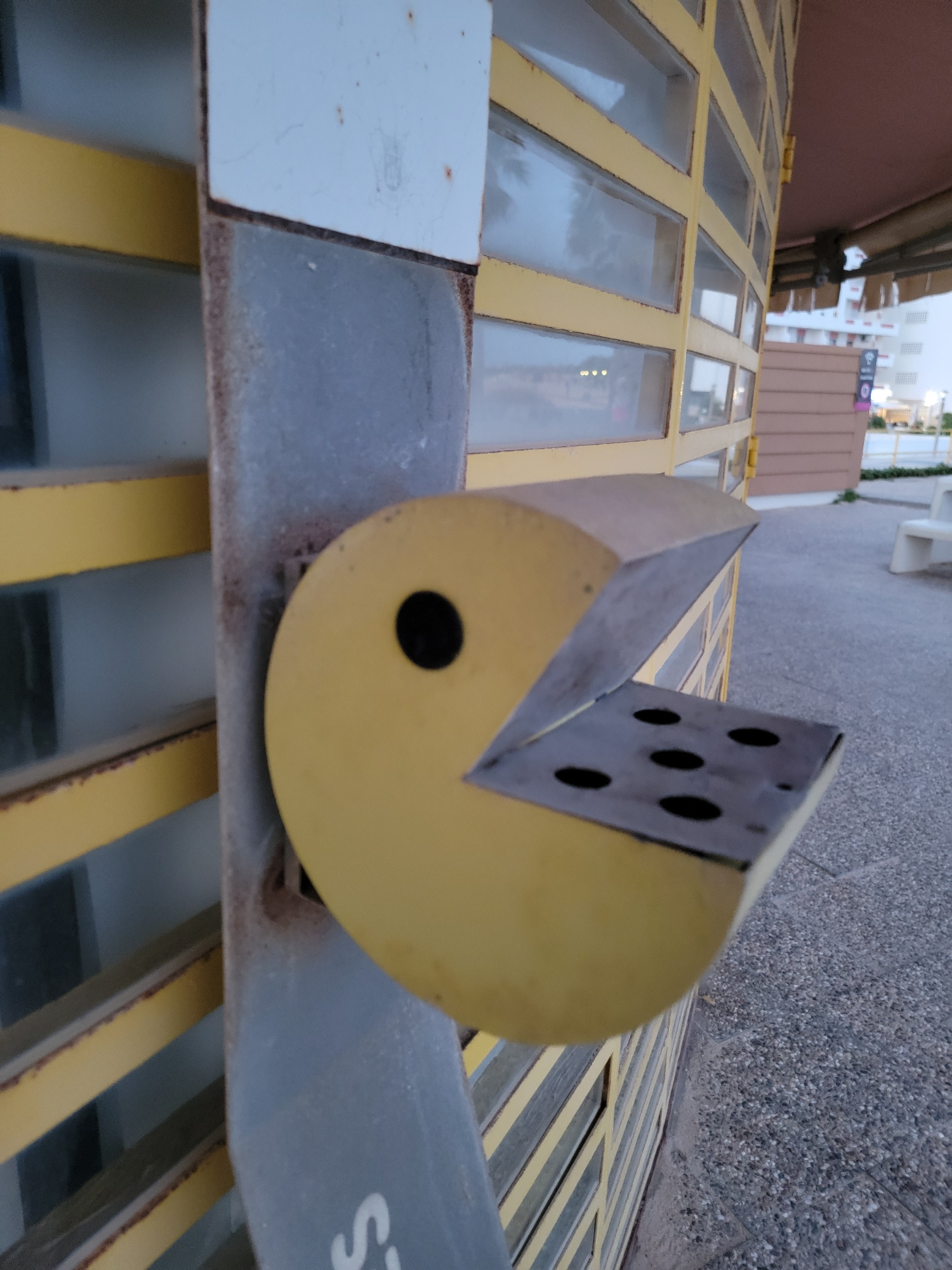 An outdoor, close-up, eye-level shot captures a yellow, Pac-Man-shaped object mounted on a grey metal pole. The object is positioned to the right of the pole, with its circular body facing left, featuring a black circular eye. The Pac-Man's mouth is open, revealing a metal tray with five circular openings. Behind the Pac-Man, a yellow roll-up door with glass panels is visible, suggesting a commercial establishment. The ground is a light grey gravel surface. In the background, a brown building and white structures, possibly buildings or apartments, are discernible, hinting at an urban or resort environment. The lighting suggests it is either early morning or late evening. The location is Quarteira, Portugal, a coastal city known for its beaches and resorts.