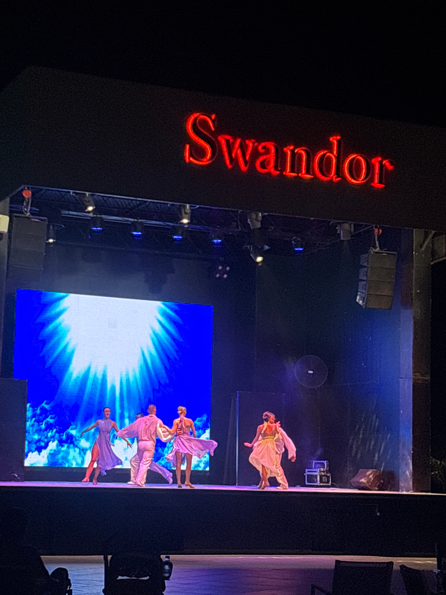 This is a photograph taken from a slightly elevated, angled perspective, capturing a performance on a stage. The scene is dimly lit, with the primary light sources emanating from stage lighting and a large screen at the back of the stage.
The prominent subject on the left is a large, illuminated sign displaying the word "Swandor" in bold, red, neon lettering. The letters are vertically oriented and stand out against a dark background.
The stage itself appears to be set for a dance performance. Several individuals, dressed in flowing, light-colored costumes, are captured mid-movement on the stage. One dancer in a light pink outfit is positioned higher up, while others in various shades of purple and pink are grouped closer together. They are performing on a stage illuminated by spotlights and a large screen at the rear. This screen displays a vibrant blue graphic, possibly abstract clouds or an explosion of light, with a bright white center radiating outwards.
Visible objects on stage include several rectangular speakers stacked in a line to the left of the screen and what appear to be lighting rigs above. There are also some stage props, including what looks like a wooden crate. The overall atmosphere suggests an evening performance. The focus is on the performance and the branding, with the dark surrounding areas indicating a theater or performance hall.