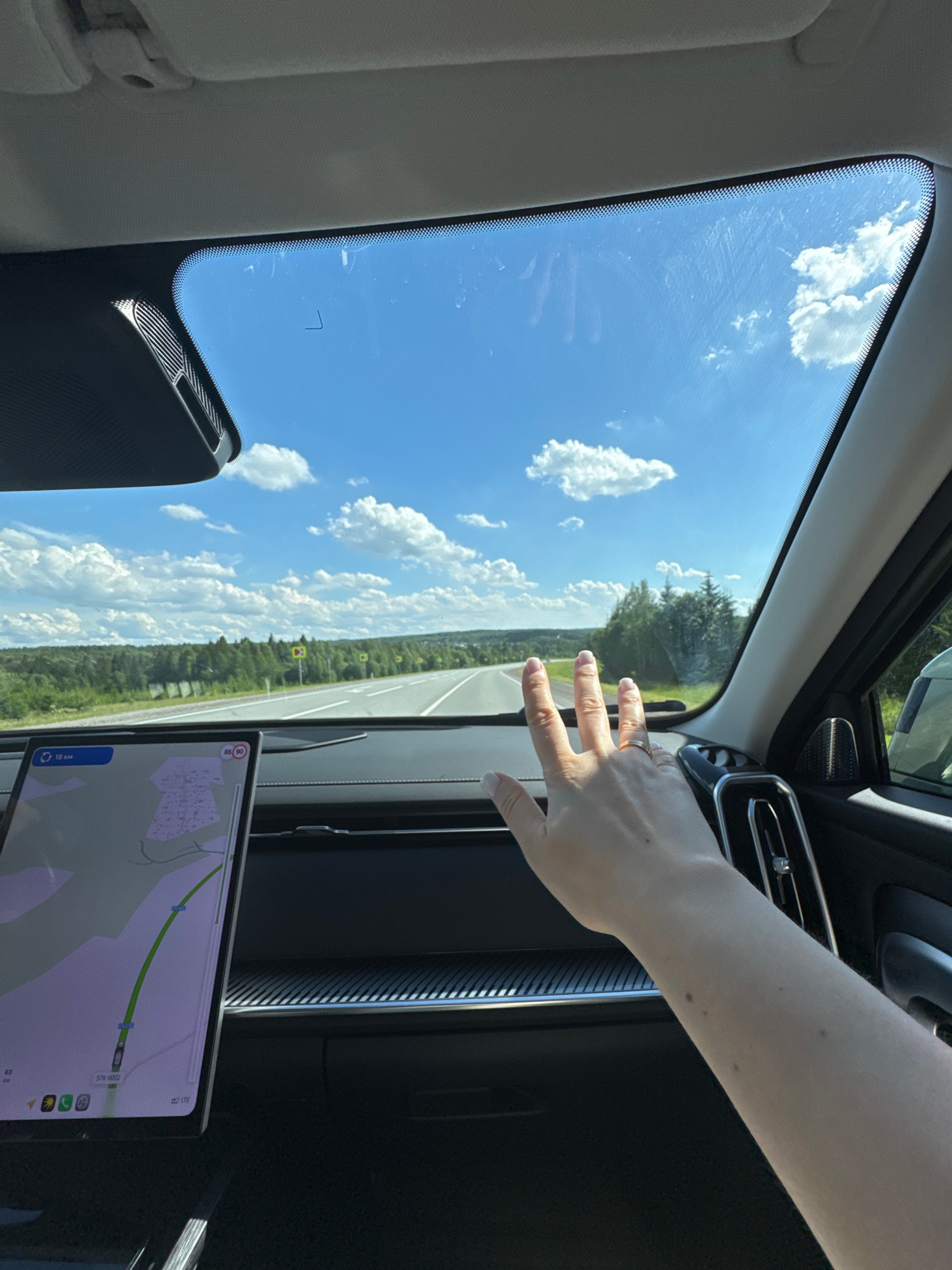 The image captures a view from inside a car, looking through the windshield. The perspective is slightly angled, with the majority of the frame filled by the bright blue sky dotted with fluffy white clouds. Trees and a distant landscape are visible through the glass. In the foreground, a person's hand with manicured fingernails and a ring on one finger is reaching out towards the windshield, seemingly interacting with the view or adjusting something. The interior of the car is visible, including a part of the dashboard with a navigation screen displaying a map. The time of day appears to be daytime, with bright sunlight. The weather seems clear and pleasant. Some faint smudges or streaks are visible on the windshield. Text is visible on the navigation screen, including "LTE", "57R-0002", and numerical values like "63" and "10 kms", indicating navigation data. The overall scene suggests a car journey through a rural or suburban area on a sunny day.