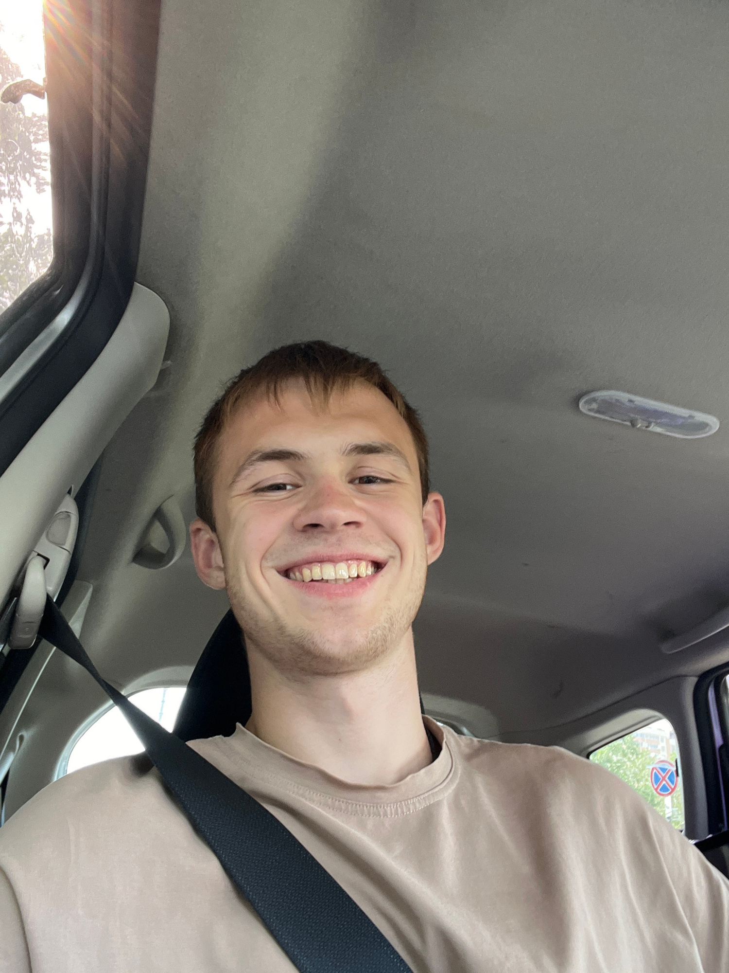 This media file is a portrait-oriented, slightly angled selfie taken from inside a car. The primary subject is a young man with short brown hair, a light stubble on his chin, and a wide, genuine smile. He is wearing a plain, light brown t-shirt and a black seatbelt is prominently visible across his chest. The background of the image shows the interior of the car, including the gray headliner and part of the dashboard. A circular, circular traffic sign with a red border, a blue background, and a red diagonal line crossing it is visible through the car window. Sunlight is streaming in from the left side of the frame, casting a warm glow and creating lens flare. The overall impression is a casual, happy moment captured while traveling. The lighting suggests it is daytime, possibly late afternoon due to the warm sunlight. No visible text is present in the image.
