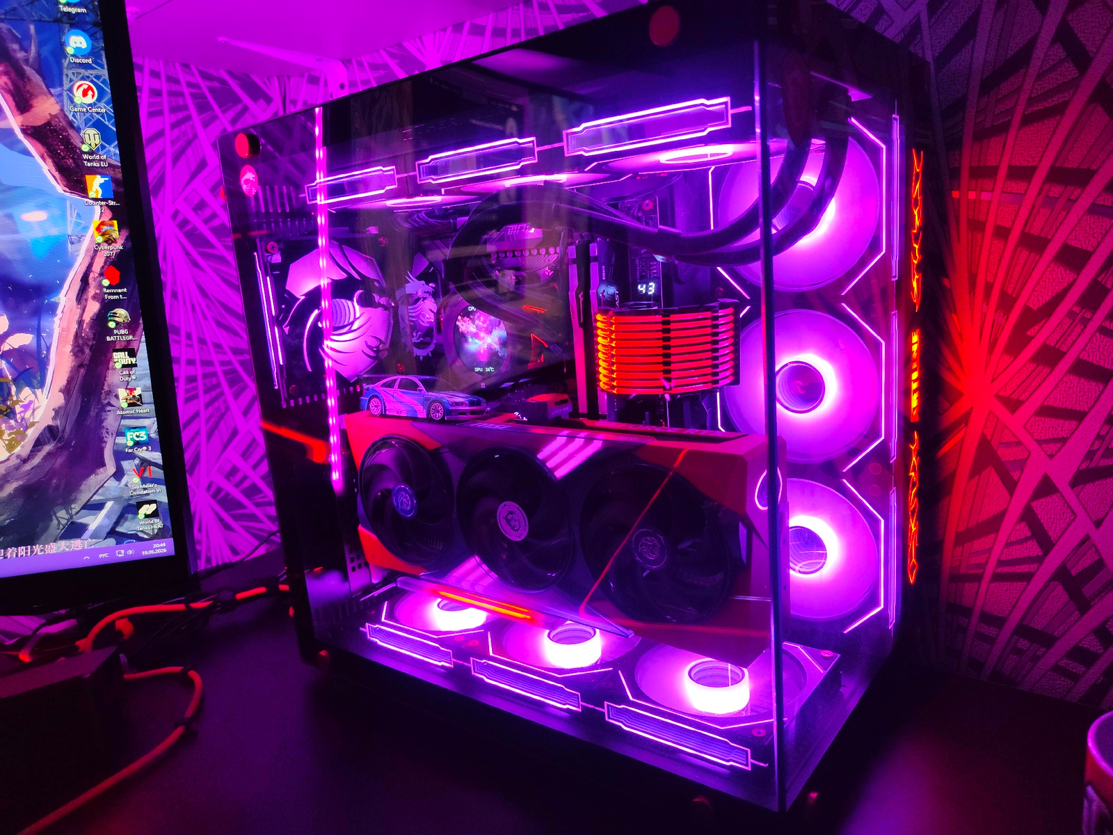 The image displays a high-end gaming computer setup, illuminated by vibrant purple and red LED lighting. The computer tower, a prominent subject, features a transparent side panel showcasing its internal components. These include a large graphics card with multiple fans, what appears to be a CPU cooler with a display, and several case fans. The tower is adorned with additional RGB lighting strips along its edges and interior. A monitor is visible to the left, displaying a desktop background with several application icons, including "Telegram", "Discord", "Game Center", and various game titles like "World of Tanks EU", "Cyberpunk 2077", and "Call of Duty". Cables, likely for the peripherals and power, are visible connecting to the computer. The environment appears to be a desk or table surface, with a patterned wallpaper or backdrop behind the computer tower. The lighting creates a distinct, futuristic ambiance. There is no discernible indication of the city of Borova, Ukraine, nor are there any people, actions, or interactions depicted in the image. The scene is static and focused solely on the computer equipment.