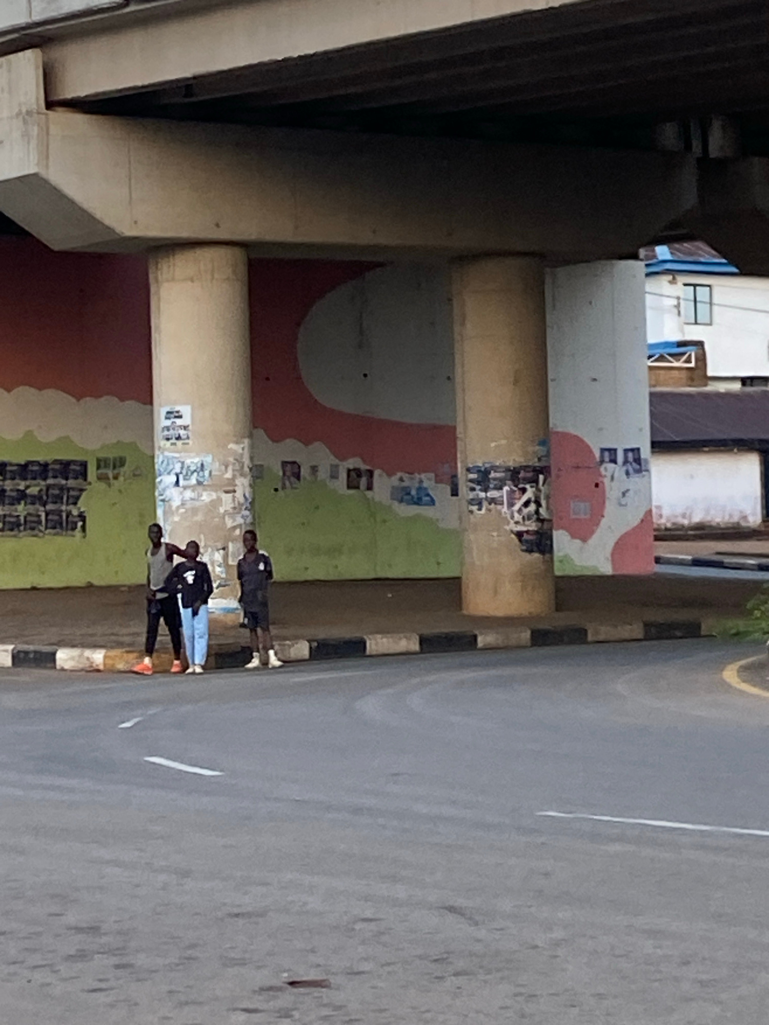 The image depicts an outdoor scene under a concrete overpass, likely in Jos, Nigeria. Three young men are standing near the base of a large concrete pillar. The pillar is covered in various posters and graffiti. The ground beneath them is a paved road with a curb. In the background, there is a wall with a colorful mural of wavy green and red shapes. Behind the mural, buildings are visible. The lighting suggests it is daytime.