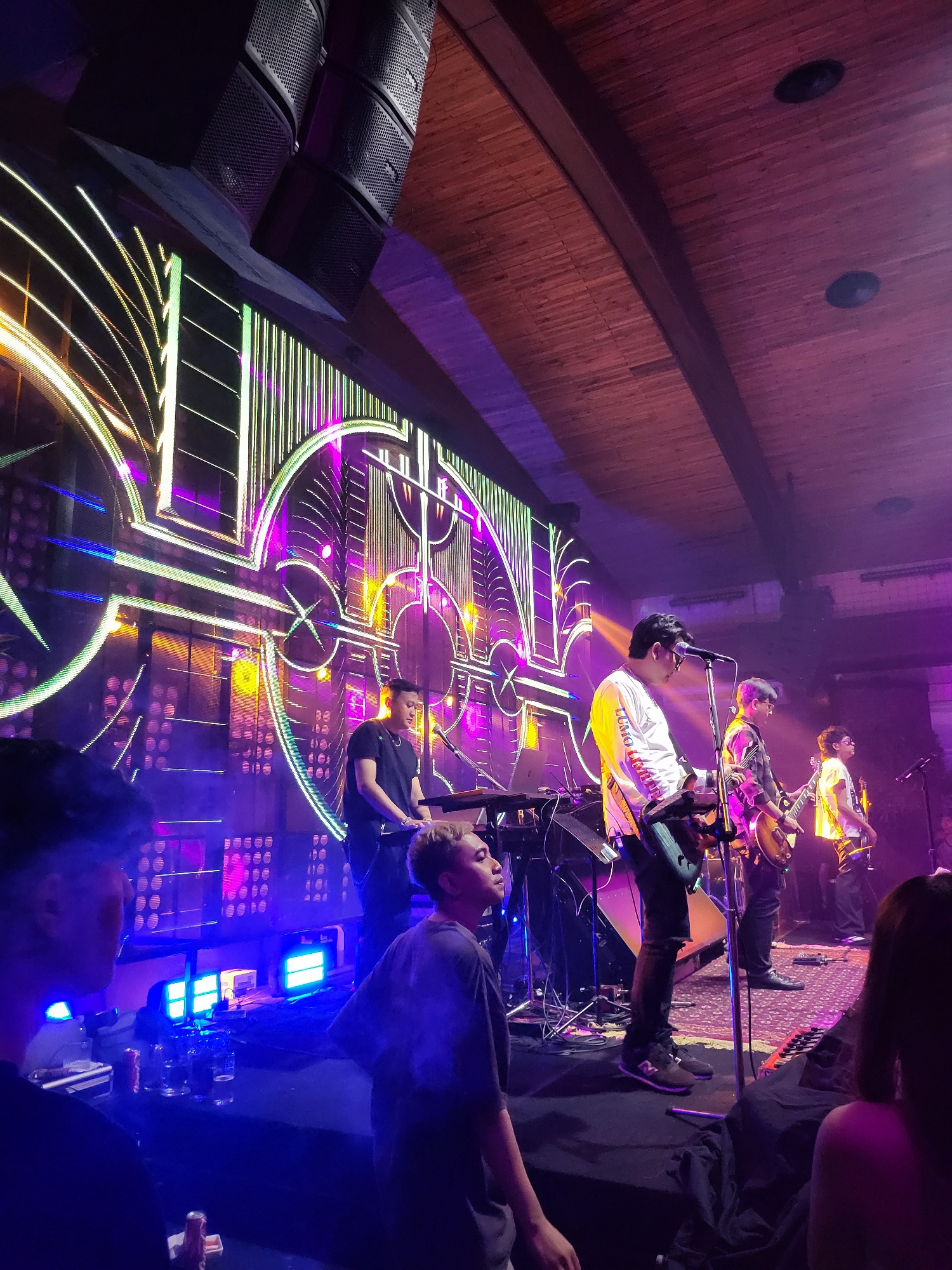 A band performs on a stage with a large LED screen behind them displaying abstract geometric patterns in neon colors of purple, yellow, and white. The stage is lit with purple and blue lights. Five male band members are visible. The lead singer in the front right is wearing a white long-sleeved shirt and dark pants, playing a keyboard. To his left, another band member in a patterned shirt plays a guitar. Further to the left, a band member in a black t-shirt stands behind a keyboard. Two other band members are partially visible on the right side of the stage, one playing a guitar and another with a microphone. In the foreground, several audience members are visible, with their backs to the camera, looking towards the stage. The room has a wooden ceiling with exposed beams. The setting appears to be a live music venue or concert hall in North Jakarta, Indonesia, likely on a weekend evening.