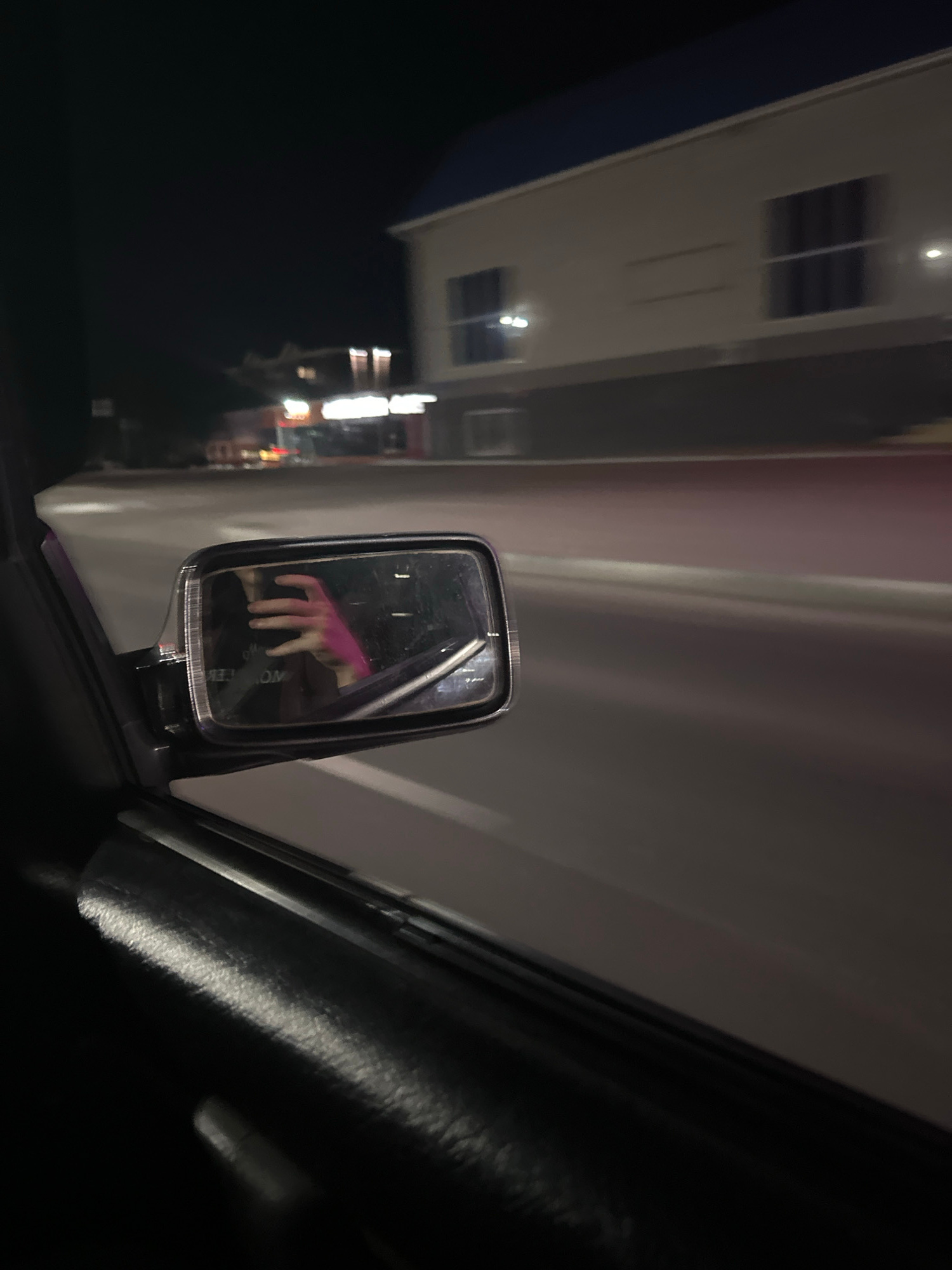 The image shows a reflection in a car's side mirror. The reflection captures a hand with pink nail polish and a black shirt. The background of the reflection is dark, with some blurred lights suggesting an urban environment at night. In the foreground, outside the mirror, is the interior of a car with a dark dashboard and a white or light-colored exterior of a building. The building appears to have windows and lights, indicating it might be a commercial establishment or residential building. The overall scene suggests the photo was taken from inside a vehicle, possibly parked or moving, at night. The presence of a hand with bright pink nail polish could imply a person is in the passenger seat or the driver is reaching out. The lighting conditions and the blurred background point to an evening or nighttime setting. There is no visible text in the image.
