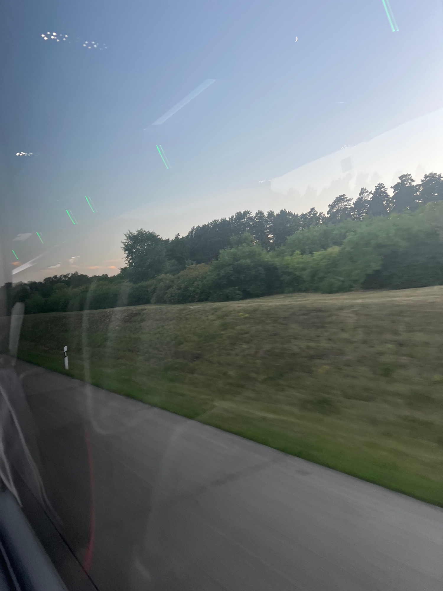 The image captures a view from inside a vehicle, likely a bus or train, looking out a window. The foreground features a blurred asphalt road with a green grassy shoulder. A white and black road marker is visible along the side of the road. Beyond the road, a slope covered in dry grass leads up to a line of lush green trees and dense foliage, suggesting a rural or semi-rural environment. The sky is a pale blue with a sliver of a crescent moon visible in the upper right quadrant. Reflections on the window obscure parts of the scene, showing streaks of light and blurry shapes, including what appear to be distant lights and linear green light sources. The overall impression is one of travel through a landscape during twilight or early morning. The location is identified as Kleinmachnow, Germany.