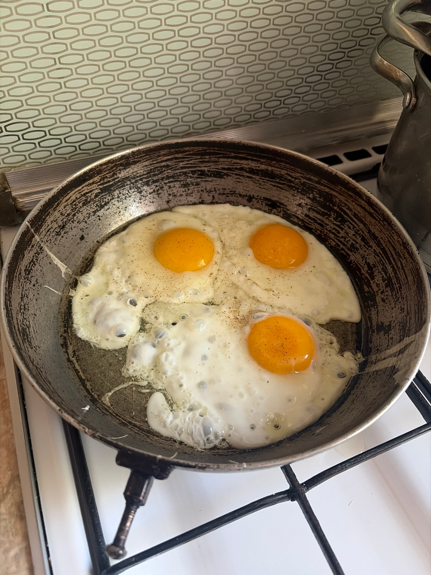 In an Unknown, Unknown kitchen, three fried eggs are cooking in a well-worn, round, metal frying pan. The pan is situated on a white gas stovetop with black grates. The eggs have bright yellow yolks and cooked white, with some bubbling in the whites. A dark-colored speckled pattern is visible on the wall behind the stovetop, and a metal pot handle is partially visible to the upper right of the frame.