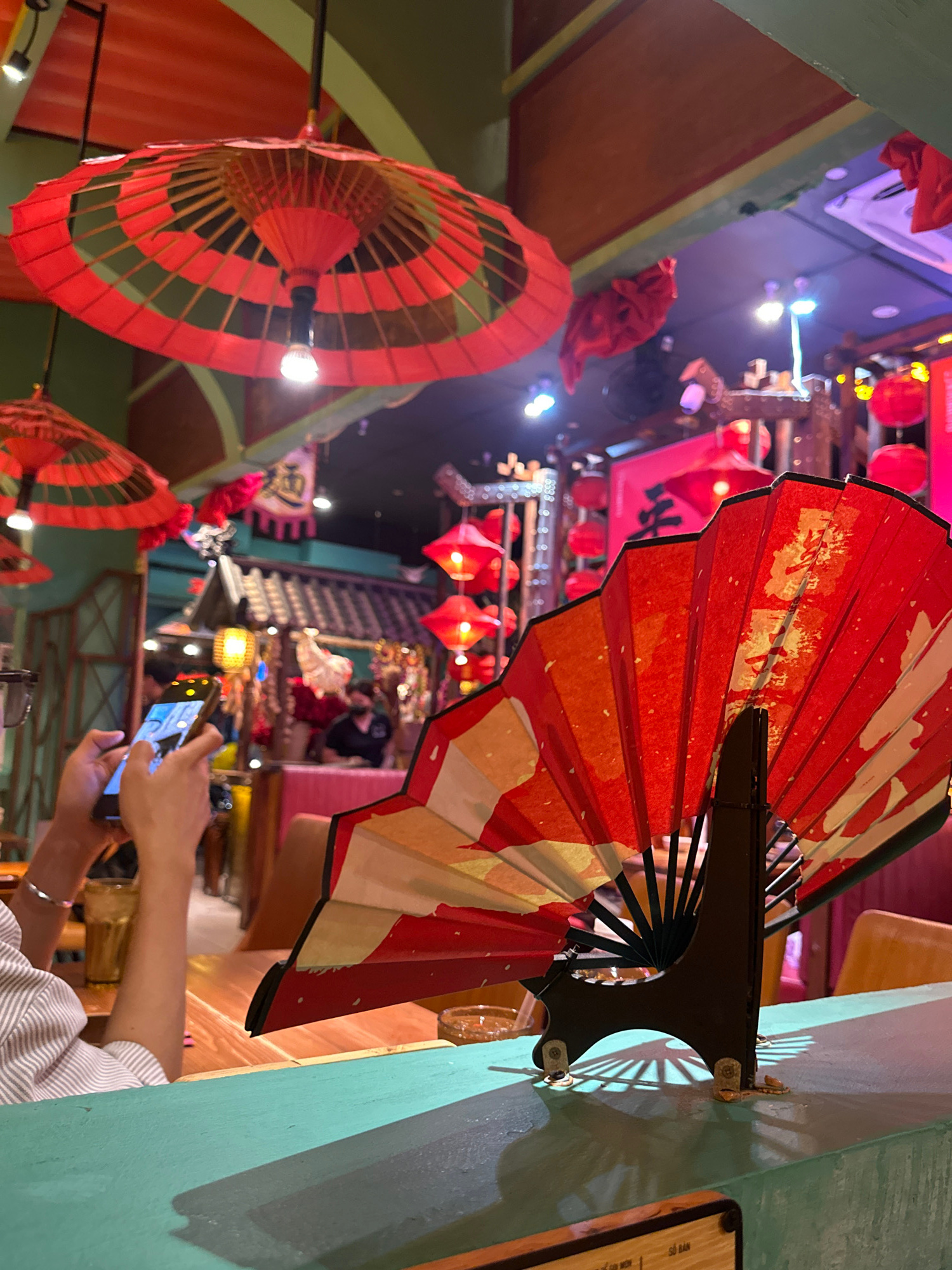 This image captures a vibrant and festively decorated interior, likely a restaurant or shop, with a strong East Asian influence. Red paper umbrellas and a large, open red and gold fan are prominently displayed, suggesting a celebratory or cultural theme, possibly related to Lunar New Year or a similar festival. Red paper lanterns also dot the ceiling, adding to the ambiance.
In the foreground, a person is holding a smartphone and appears to be taking a picture or video of the scene. Their hands are visible, and they are wearing a striped shirt. The perspective is from behind and to the side of this individual, looking into the space.
The setting is dimly lit with warm lighting, creating a cozy and inviting atmosphere. Various levels and structures within the space are visible, hinting at a multi-tiered design. The walls are painted in a contrasting teal color.
While the exact time of day isn't specified, the indoor lighting suggests it could be evening or night, or a dimly lit indoor environment regardless of the external time. There are no clear indications of weather.
A small piece of paper with some text is visible on the right side of the frame, but the text is largely unreadable due to its size and the angle. However, some Vietnamese characters are discernible, hinting at the location being in Vietnam.
The overall impression is one of a lively, culturally rich environment, captured in a moment by a visitor or patron.