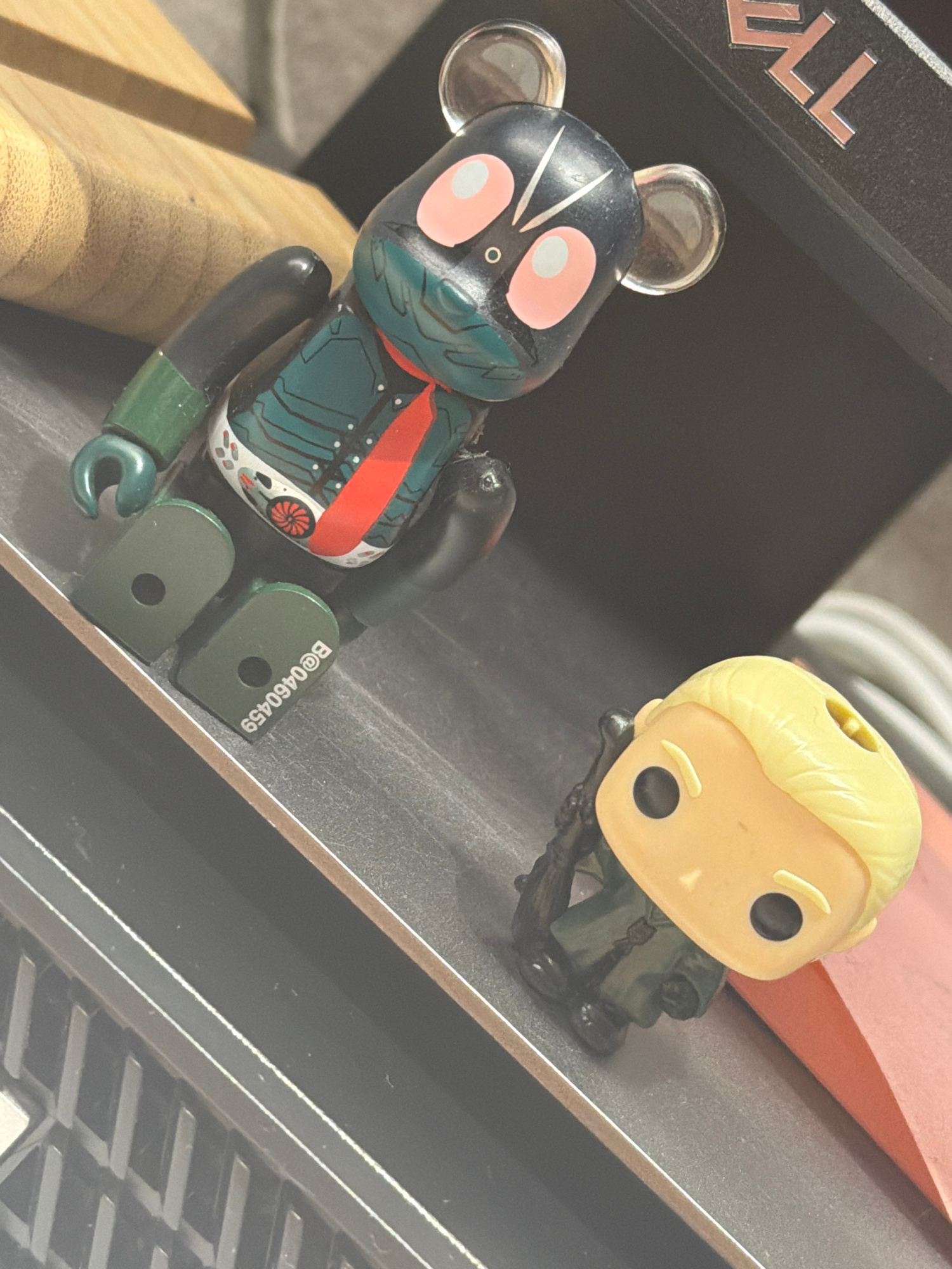 Two collectible figurines are placed on a dark, textured surface. The figurine on the left is a black bear-shaped toy with pink circular eyes and green accents on its limbs and torso, featuring a red tie and a white and green patterned chest. The figurine on the right is a smaller, yellow-headed figure with black eyes and blond hair, dressed in a dark green military-style outfit and holding a rifle. In the background, a wooden object is partially visible on the left, and a black object with metallic letters that appear to spell "TELL" is present on the upper right. The scene appears to be indoors, possibly on a desk or shelf, with no clear indicators of its specific location within Almaty, Kazakhstan.