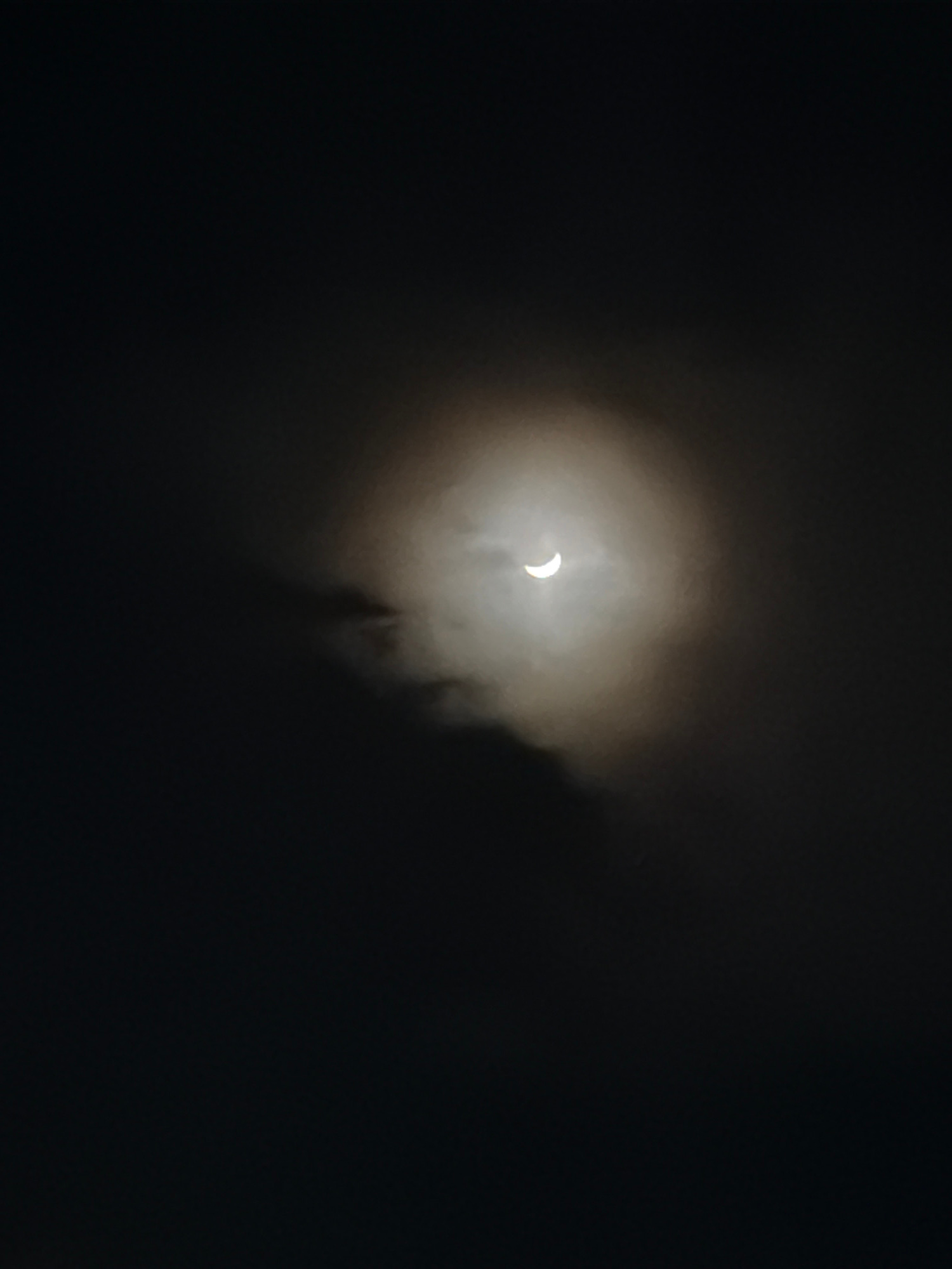 The image depicts a partial solar eclipse observed through clouds. The sun, partially obscured, appears as a crescent shape surrounded by a hazy glow. The sky is predominantly dark, suggesting either nighttime or heavy cloud cover. There are no visible people, specific objects, or distinct landscape features that would clearly indicate a location, other than the provided context of Choeng Mon, Thailand. The overall mood is somewhat mysterious due to the low visibility and the celestial event. No text is present in the image.