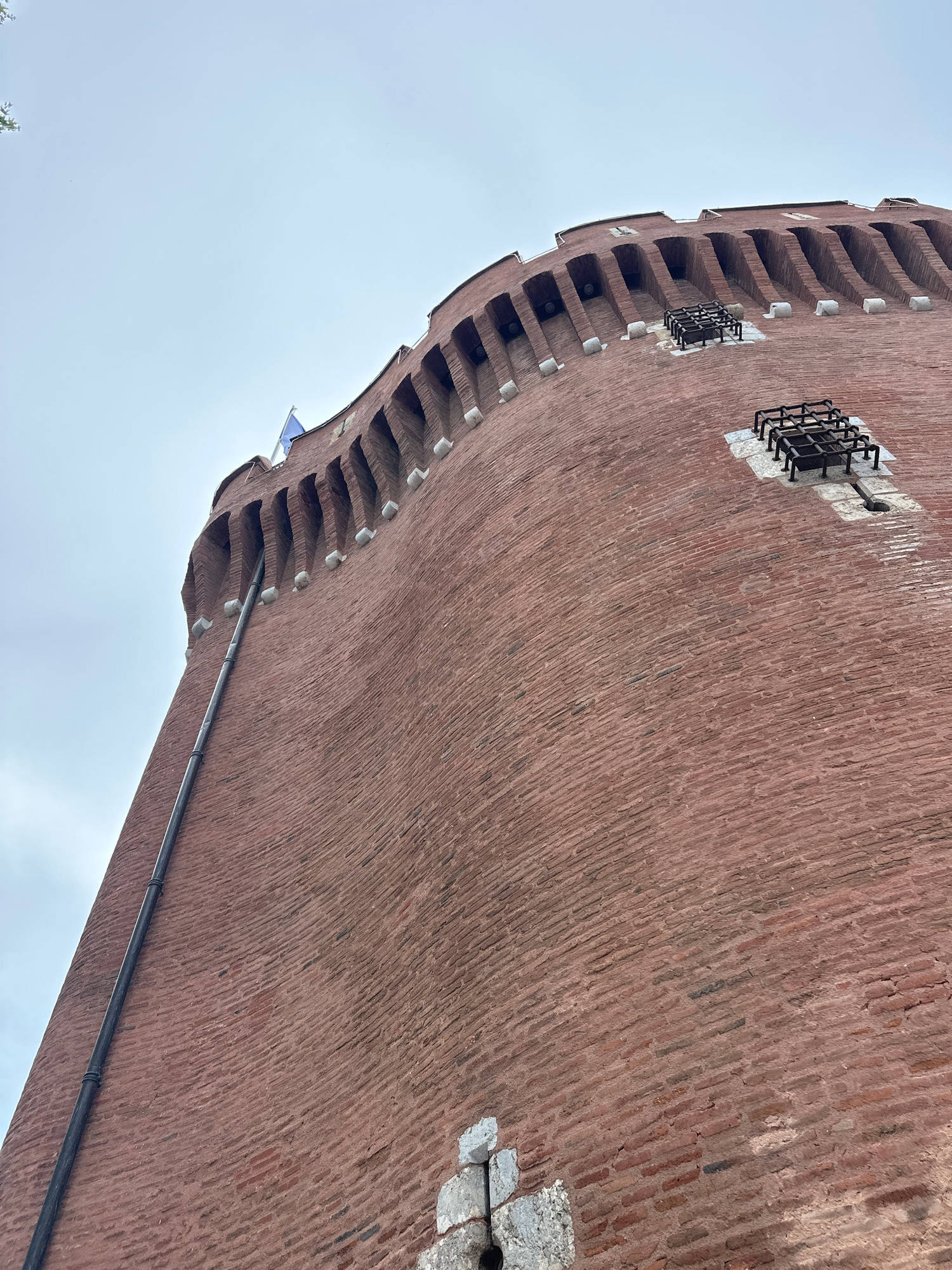 The image shows a low-angle, upward view of the exterior of a tall, circular, red brick tower in Perpignan, France. The tower has a crenellated top with arched openings, and a dark pipe runs vertically along its side. Two square, metal grate structures are visible on the brick surface. A portion of a blue flag is visible at the top of the tower. The sky is overcast and pale. The context provided indicates this is likely a section of the Castillet, a historic monument and former city gate in Perpignan.