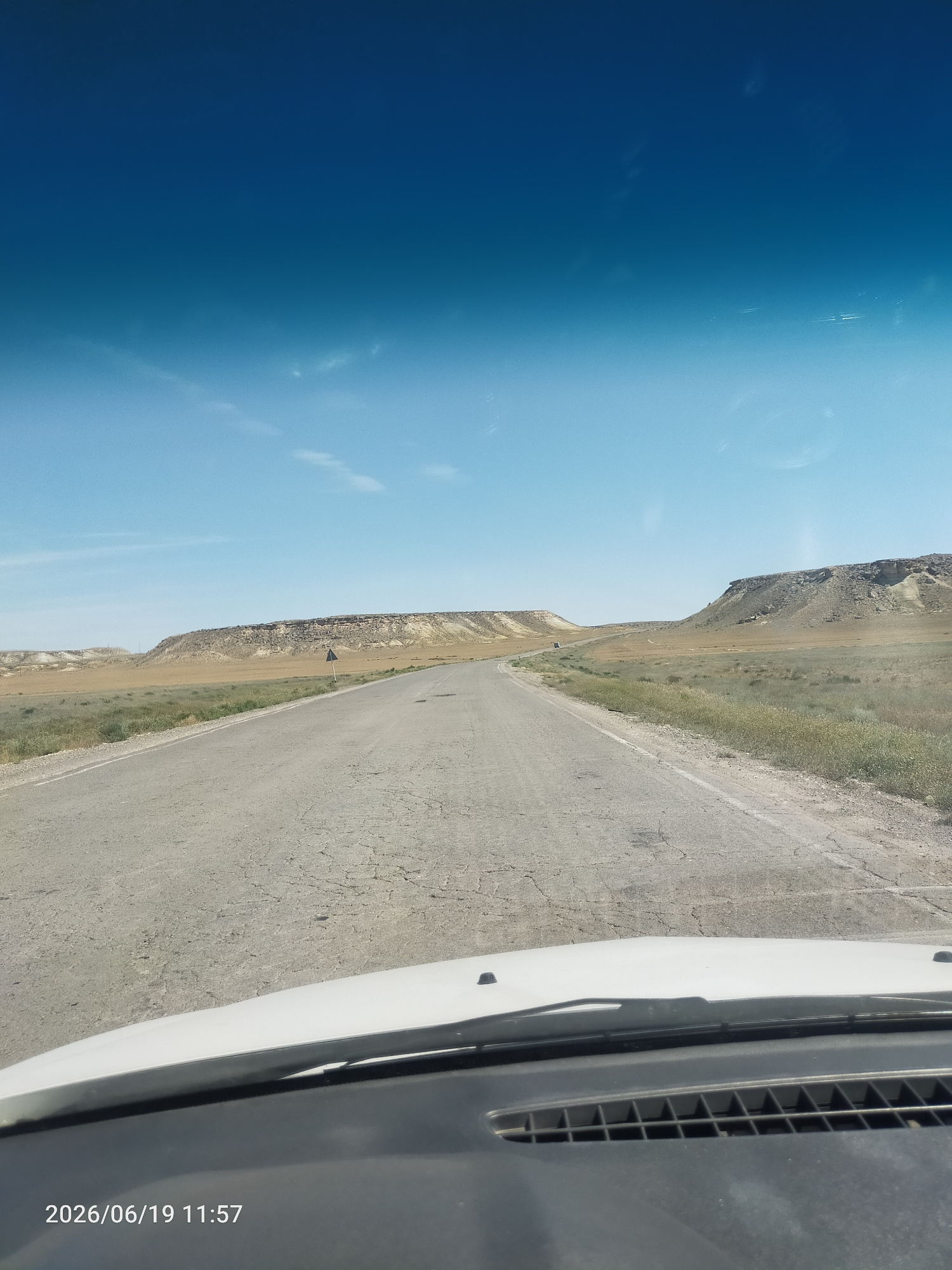The image captures a view from inside a vehicle, looking out at a desolate, sun-drenched landscape. The foreground is dominated by the white hood and black dashboard of the car, with a portion of the windshield visible. The road ahead is cracked and appears to be asphalt, stretching into the distance. On either side of the road, dry, sparse vegetation grows, giving way to gently rolling hills and plateaus in the background. These geological formations are layered with varying shades of tan, brown, and white, suggesting a dry, arid environment. The sky is a clear, vibrant blue, with a few wispy clouds scattered across it. The sun is high, indicating it is midday. A solitary triangular road sign, black with a white border, is visible to the right of the road in the middle distance. The overall impression is one of vastness and isolation. The date and time stamp at the bottom left corner reads "2026/06/19 11:57".