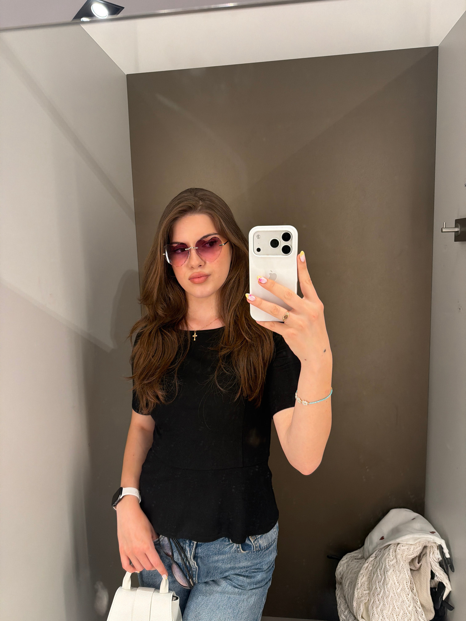 A young woman with long brown hair and pink heart-shaped sunglasses takes a mirror selfie in a fitting room. She is wearing a black peplum top, blue jeans, a white smartwatch, and a delicate gold cross necklace. Her right hand, holding an iPhone, is adorned with a ring and a beaded bracelet. Her nails are painted with a pastel yellow and pink design. A small white handbag is held in her left hand. The fitting room has grey walls and a dark brown background. A pile of folded clothing, including a cream-colored knit sweater, rests on a shelf in the corner. The image was captured in Sofia, Bulgaria.