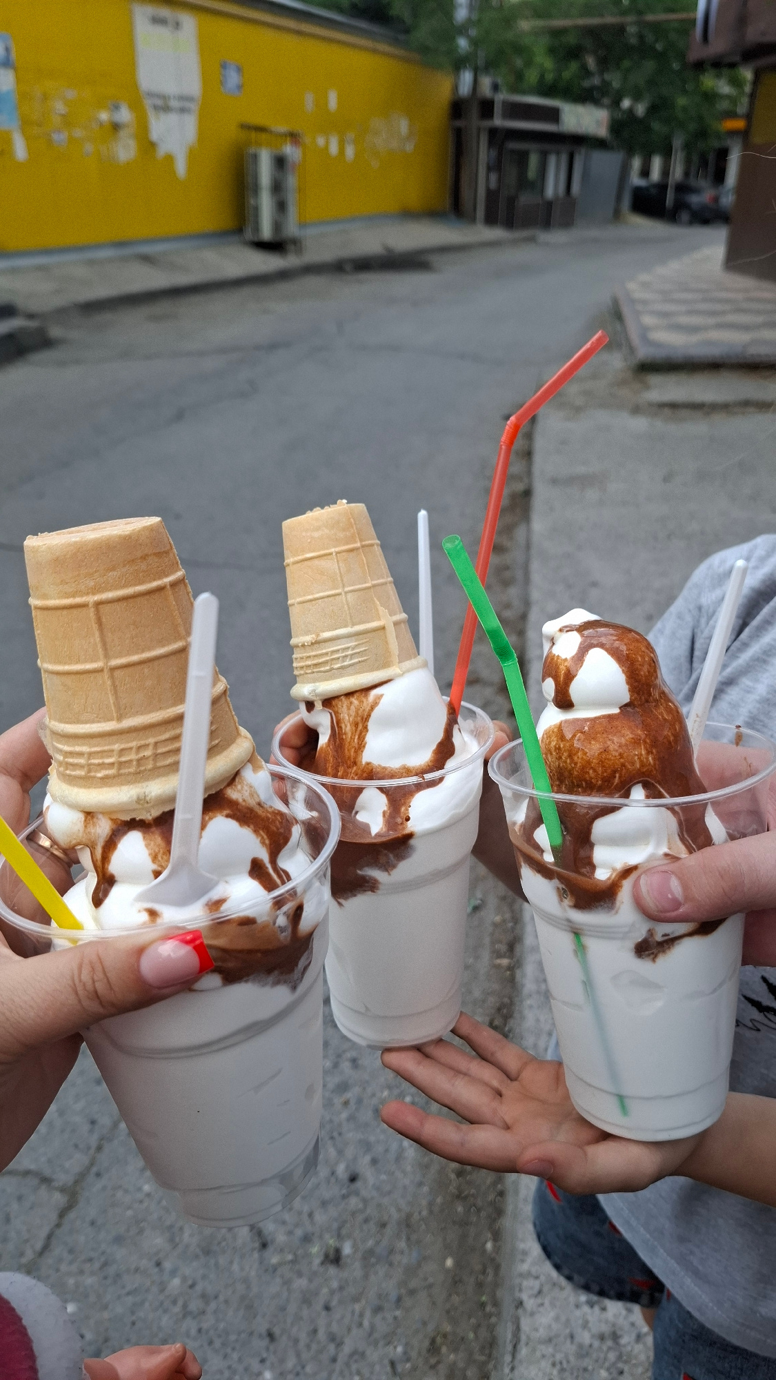 Three cups of ice cream are held by two people, with two cups being held from the left and one from the right, and a child's hand supports the middle cup. The ice cream in the cups is white with chocolate syrup drizzled on top. Two of the ice cream cups have waffle cones inverted and placed on top. Each cup has a straw. The background shows a street scene with a yellow building on the left, suggesting an urban environment. The presence of a yellow building and the overall streetscape are consistent with an outdoor location in the city of Shymkent, Kazakhstan, possibly near a street vendor or an ice cream shop.