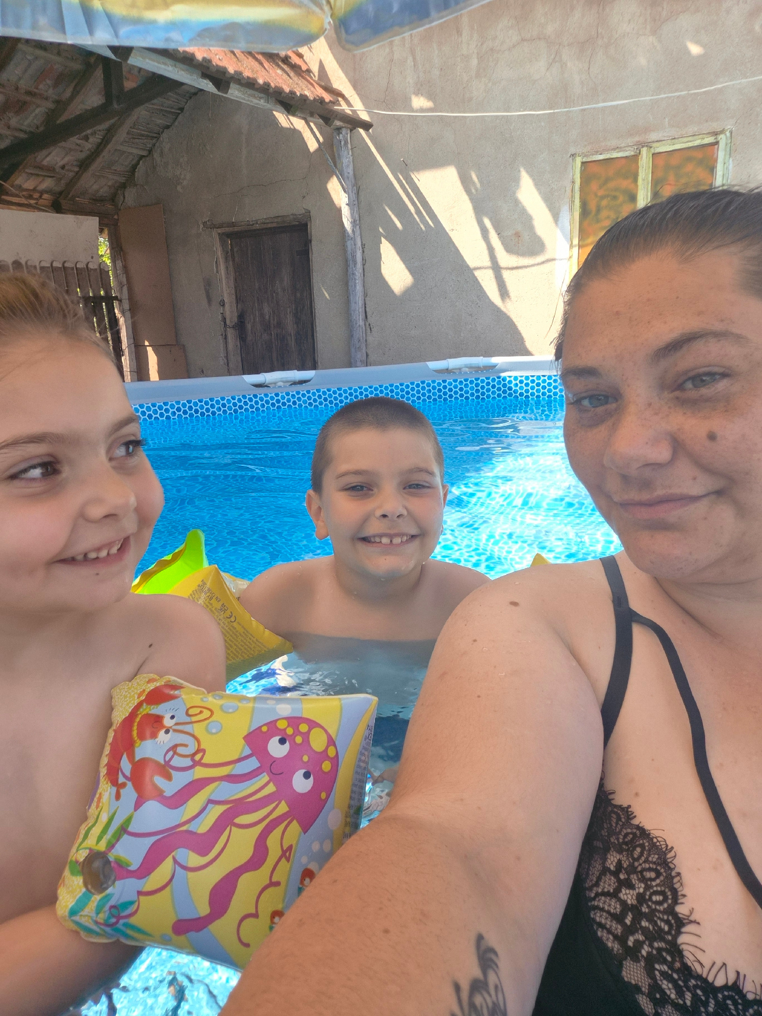 In an outdoor swimming pool, a woman and two children are enjoying a sunny day. The woman, with freckles across her face and a black lace bikini top, is in the foreground, smiling at the camera. To her left and slightly behind her, a young boy, also smiling, is visible in the pool. Further back and to the left, a girl with bright eyes and a wide smile is seen, wearing yellow arm floaties adorned with a pink octopus design. The pool water is a vibrant blue. Behind the pool, a rustic building with a weathered wooden roof and walls is visible, hinting at a rural or older setting. The time of day appears to be daytime, with strong sunlight casting shadows on the building. The overall atmosphere is one of joy and family fun. Visible text on the arm floaties includes "CE EN 13138-1". The location is Kalipetrovo, Bulgaria.