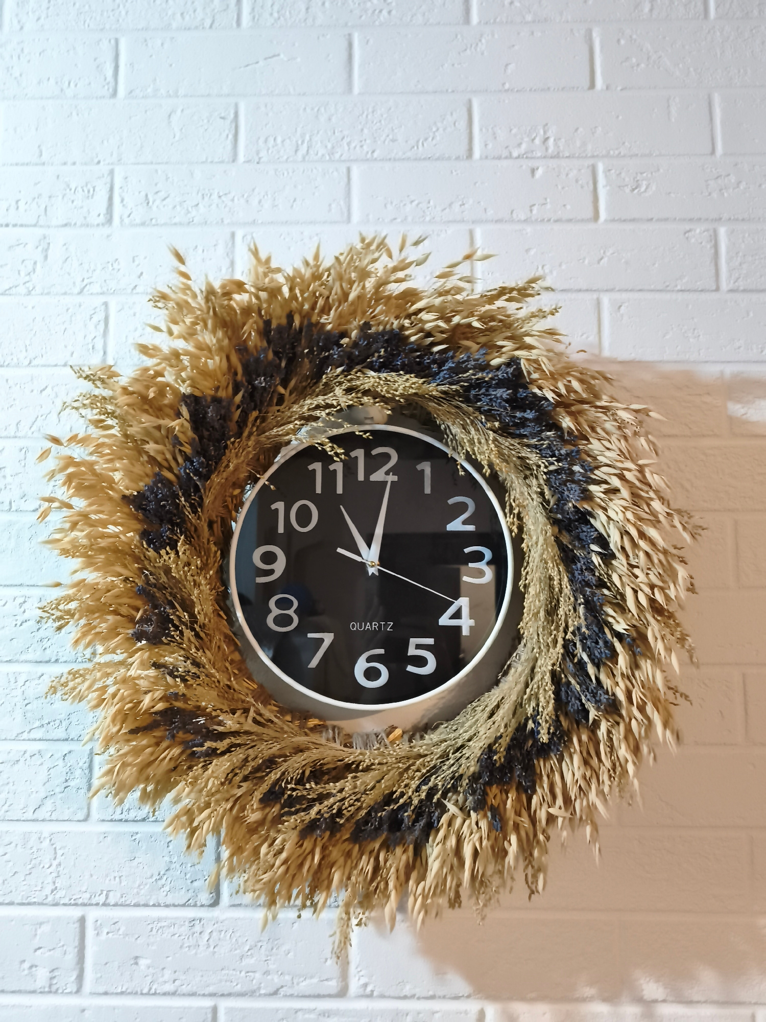 A round, black-faced clock with white numbers and hands is centered on a white brick wall. The clock is surrounded by a decorative wreath made of dried wheat stalks and dark purple sprigs, possibly lavender. The wreath is artfully arranged to frame the clock face. The lighting suggests an indoor setting, with the white brick wall showing texture and slight shadows. The scene is static, with no people or apparent actions. The location is identified as Zadebri, Ukraine.