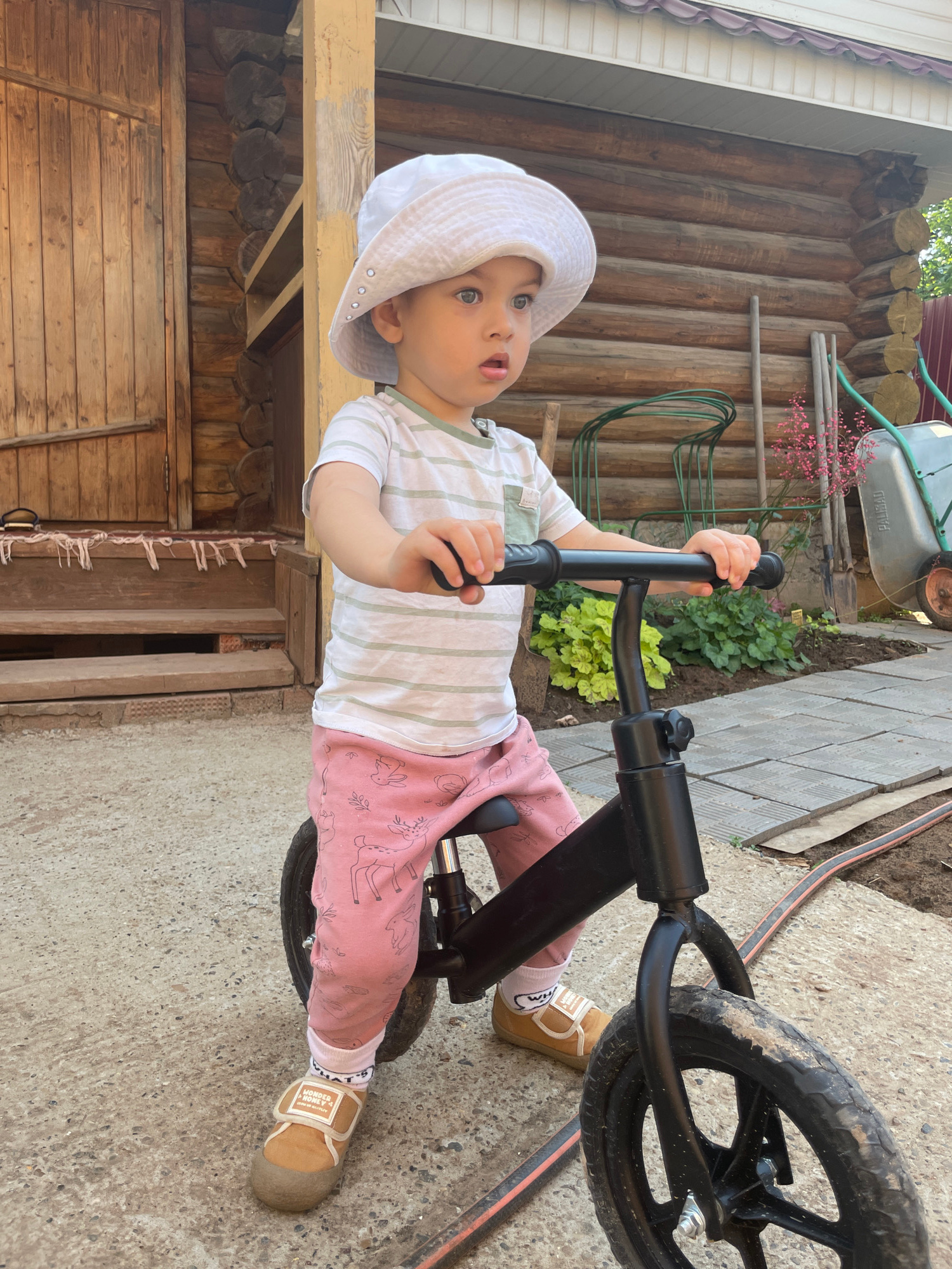 A toddler wearing a white bucket hat, a striped t-shirt, and pink pants with animal prints sits on a black balance bike. The child has blue eyes and is looking to the right with their mouth slightly open. The background shows a wooden building with a porch and steps, a garden with plants, and a wheelbarrow. The scene appears to be outdoors on a paved area. The image is taken from a low angle, focusing on the child and the bike. The location is identified as Zav'yalovo, Russia.