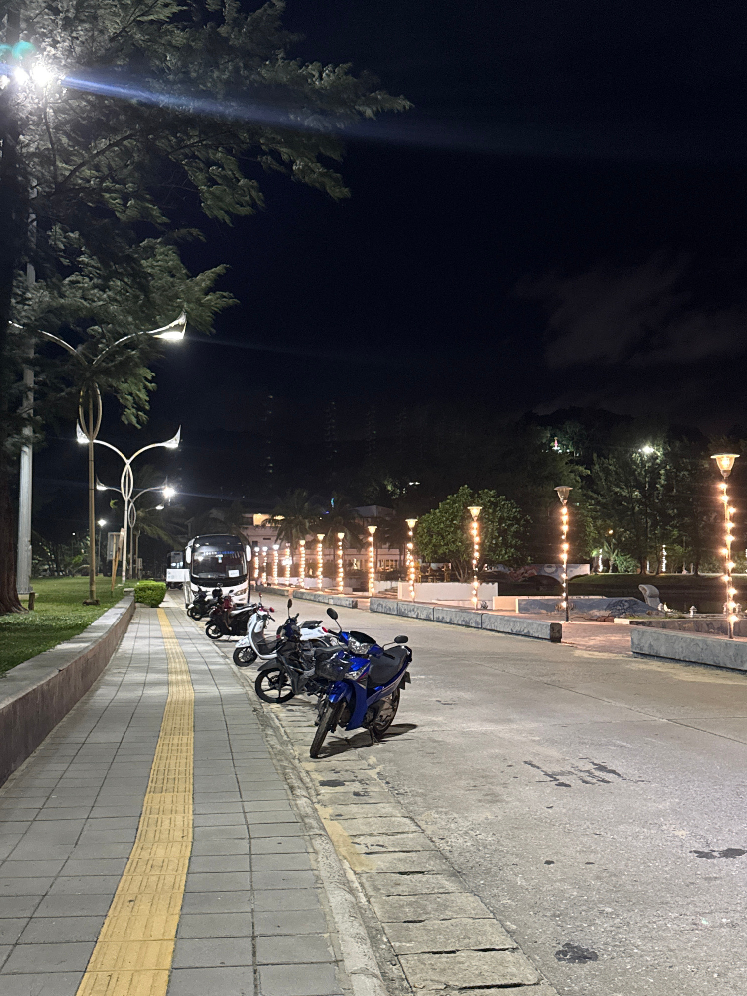 This media file captures a nighttime scene in Karon, Thailand, likely along a coastal promenade or street. The dominant subjects are several parked motorcycles, including a prominent blue and black sportbike, and a white scooter. A bus is also visible parked further down the street. The setting is an outdoor public space with a paved walkway featuring a distinctive yellow tactile paving strip, bordered by a low wall and a grassy area. Beyond the walkway, trees and palm trees are illuminated by strings of decorative lights, creating a festive atmosphere. Several streetlights with distinctive modern designs are also present. The overall mood is serene and slightly ethereal due to the night setting and artificial lighting. The time of day is clearly night, with a dark sky and artificial illumination being the primary light sources. There are no visible people in the scene. No text is legible.
