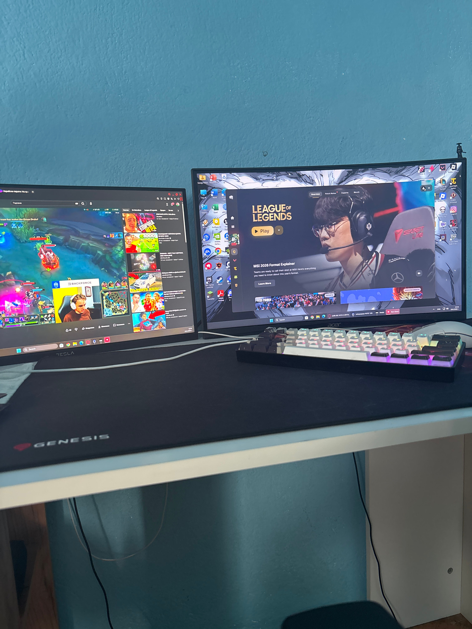 The image shows a gaming setup with two computer monitors and a keyboard. The top monitor displays the League of Legends logo and a video thumbnail of a person wearing headphones. The bottom monitor shows gameplay from League of Legends. The keyboard is white with RGB lighting. The monitors are placed on a black mousepad with the "TESLA" and "GENESIS" logos visible. The background is a blue wall. There are also some cables visible behind the monitors. The image appears to have been taken indoors, likely in a home setting. The activity taking place is gaming. The time of day and weather are not discernible. There are no people clearly visible in the image, but the content on the screens suggests a gaming context. Visible text includes "LEAGUE OF LEGENDS", "TESLA", and "GENESIS". The location context is Gabrovo, Bulgaria.