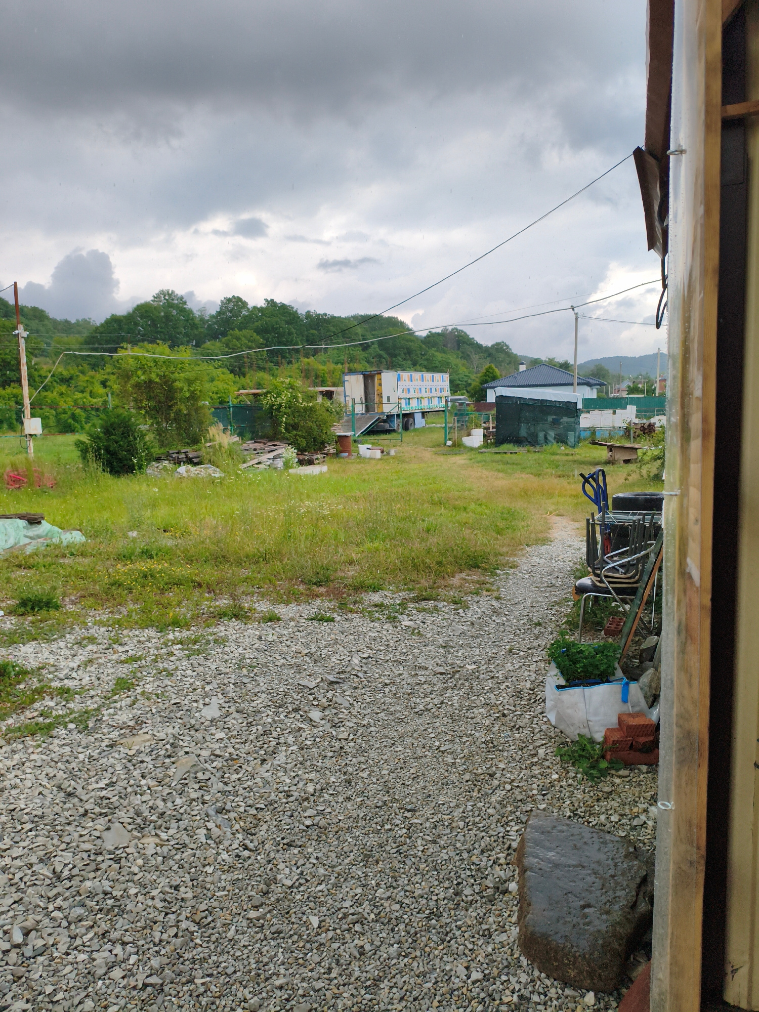 This is a medium shot of an outdoor scene in Vozrozhdeniye, Russia, under a cloudy and possibly rainy sky. The foreground is dominated by a gravel path leading into a grassy area. To the right, a wooden structure frames the right edge of the image, and some discarded objects are piled near it, including chairs, a tire, and a large rock. A white bag with green plants sits beside the objects, along with some bricks.
In the middle ground, the grassy field is scattered with various items, including pieces of wood and what appear to be construction materials or debris. Beyond the field, there are several buildings and structures. A light-colored building with a blue roof is visible, along with a taller white structure that seems to be a mobile unit with windows. Further back, a lush green, tree-covered hill rises under the overcast sky. There are electrical wires crisscrossing the scene, and what might be small raindrops are visible in the air. The overall impression is one of a somewhat unkempt rural or semi-rural area. The weather suggests it might be late afternoon or early evening, given the diffused light and the dark clouds.