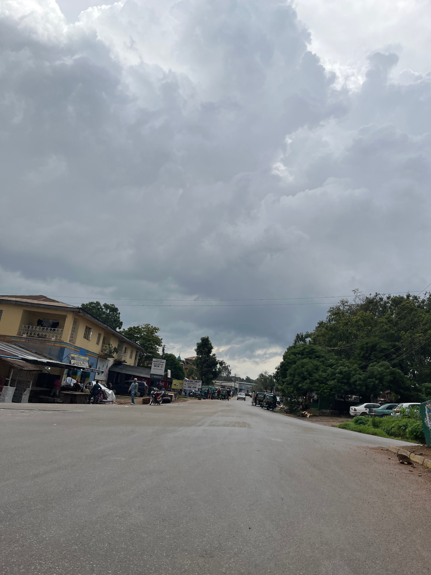 The image depicts a street scene in Jos, Nigeria, under a heavily overcast sky with dramatic cloud formations. The road, paved and appearing wet, stretches into the distance. On the left side of the road, a two-story building with a yellow facade and balconies stands, adjacent to a row of shops with corrugated iron roofs. Signs for "INFINITY FURNITURE" are visible. Several people are gathered in front of these shops, some seated. A few motorcycles and a car are parked nearby.
On the right side of the road, lush green trees form a dense barrier. Several vehicles, including cars and what appear to be auto-rickshaws, are parked along the roadside. The overall impression is of a moderately busy street scene, possibly on the outskirts of a town or in a less densely populated urban area, with the impending weather contributing to a subdued atmosphere.