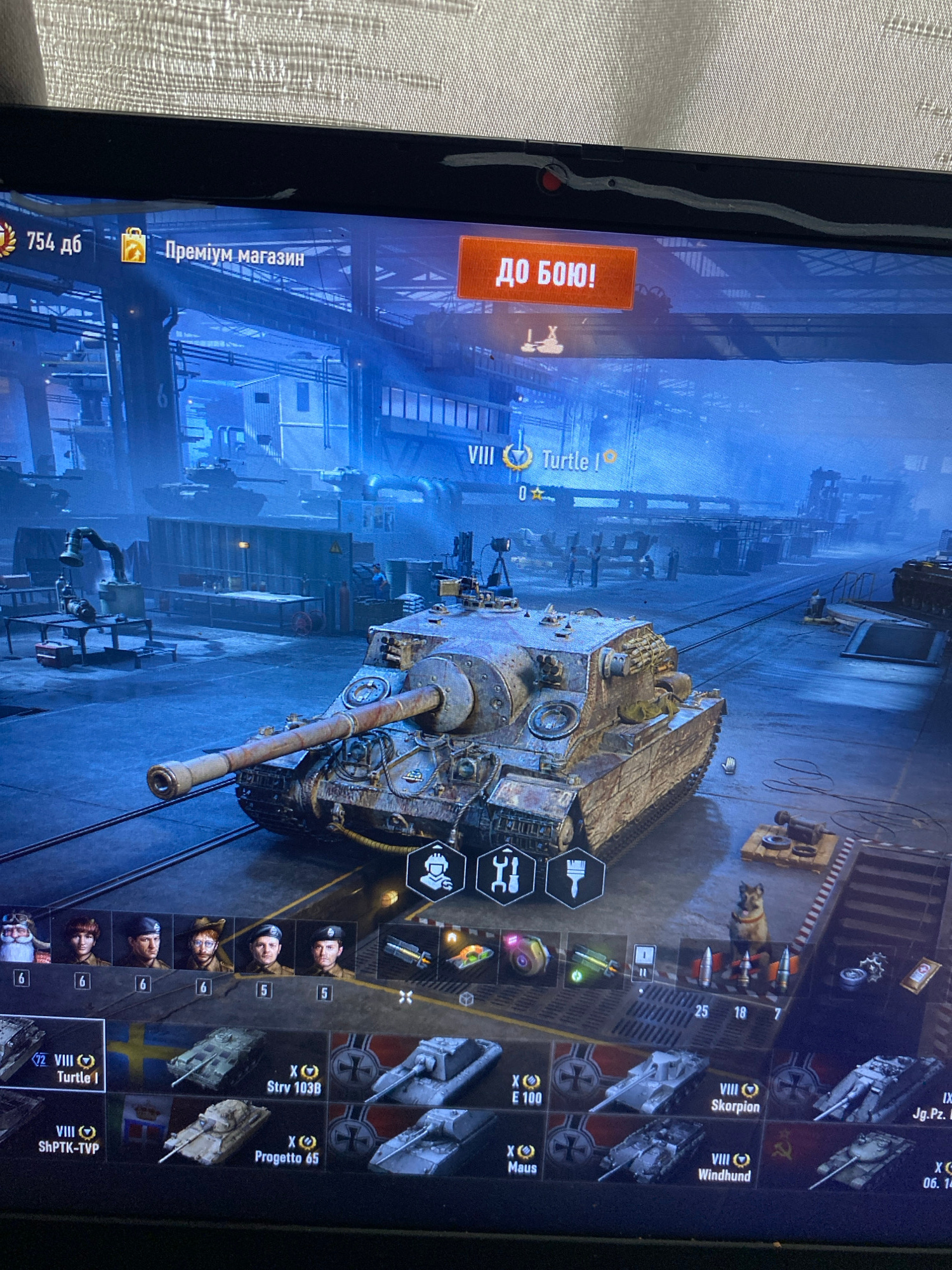 The image shows a computer screen displaying the loading screen of a video game, likely a tank combat simulator. The game interface is visible, with elements such as a "754 дб" notification (possibly in-game currency or experience points), a "Преміум магазин" (premium shop) button, and a large "ДО БОЮ!" (To battle!) button. A detailed 3D model of a tank is prominently featured in the center of the screen, appearing to be in a garage or workshop environment with other tanks and industrial elements in the background. The game's user interface also shows icons for crew members, consumable items, and available tanks in a player's inventory. The screen's reflection shows a textured surface, likely a wall or curtain. The computer is placed on a surface, the texture of which is also visible. The scene does not provide any specific information or context that would allow for the identification of the city of Szczecin, Poland.