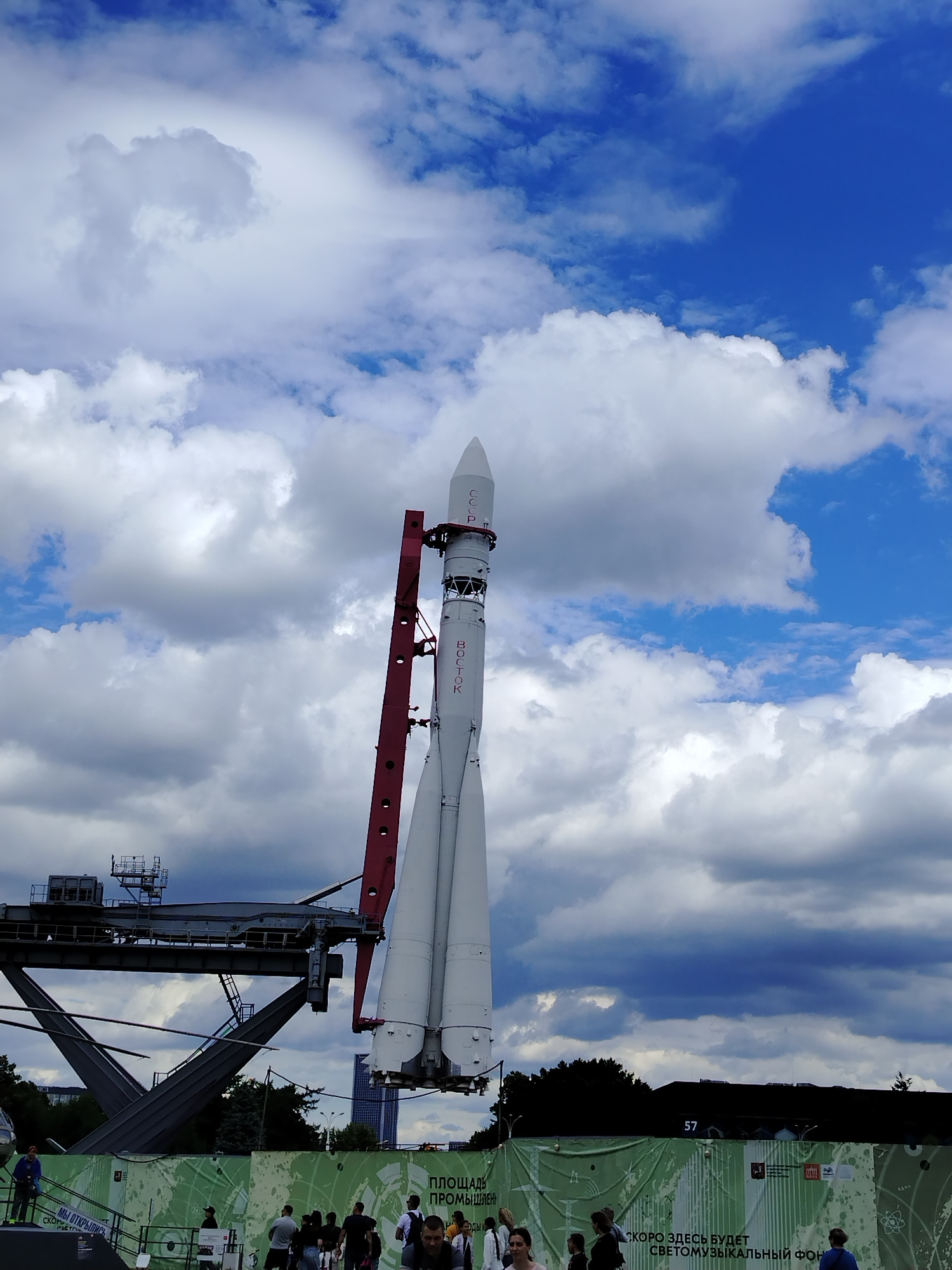 A Vostok rocket stands vertically on a launchpad against a cloudy blue sky. The rocket is white with red lettering that reads "СССР" and "ВОСТОК". A red metal structure is attached to the side of the rocket. In the background, there are trees and buildings. People are visible in the foreground, walking around the launchpad area. A green banner with text and graphics is displayed in front of the people. The scene appears to be an outdoor exhibition or museum.