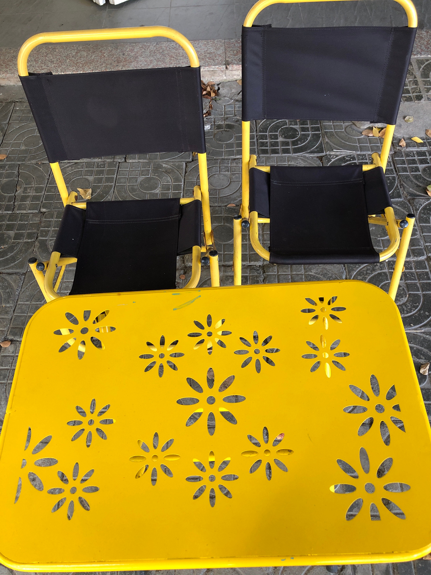 This image shows two yellow-framed black canvas chairs and a yellow metal table with a decorative floral cut-out pattern. The furniture is positioned on a grey tiled surface, with fallen leaves scattered around. The scene appears to be outdoors, possibly on a sidewalk or patio. While the location is stated to be Đà Nẵng, Vietnam, there are no specific visual cues in the image to confirm this. The image captures a static, everyday scene with no visible people or animals. The lighting suggests it is daytime.