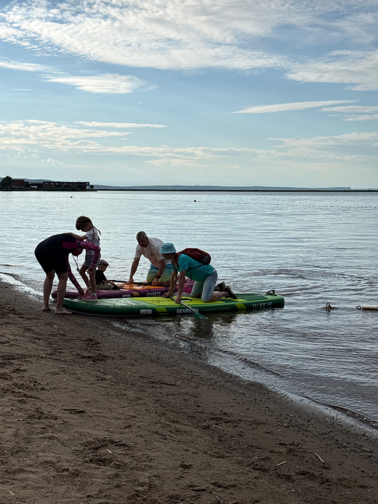 The image depicts a group of people preparing for or disembarking from paddleboarding on a body of water, likely the Kama River, near the city of Chaikovsky, Russia. The scene is set on a sandy beach with the water lapping at the shore. A variety of paddleboards are present, some partially submerged in the water and others on the sand.
In the foreground, on the left, a person in a dark full-body swimsuit with pink accents is bent over, interacting with a young girl who is standing and holding onto the swimsuit. Another child, wearing a cap, is sitting on a paddleboard in the middle. To the right of these figures, a man in a white t-shirt and shorts is kneeling on a paddleboard, and further to the right, another person in a teal shirt and shorts is also kneeling on a paddleboard. The people appear to be engaged in a group activity related to paddleboarding.
The environment is characterized by a calm, rippled body of water, a sandy beach, and a cloudy sky with patches of blue. In the distance, across the water, a shoreline is visible with some buildings and structures, possibly part of the industrial or residential area of Chaikovsky. A breakwater or barrier line is also discernible in the water, extending towards the horizon. The lighting suggests it is daytime, likely late afternoon or early evening due to the soft quality of the light.
