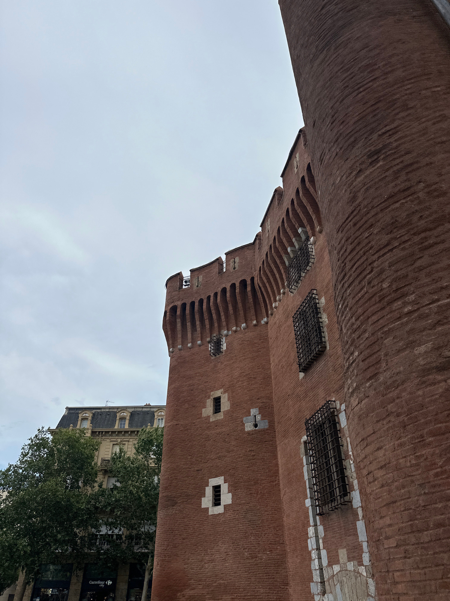 This is an image of a brick tower with distinctive architectural features, including crenellations and barred windows. The tower is made of red brick and appears to be quite old. The image is taken from a low angle, looking up at the tower, and it's rotated 90 degrees clockwise.
In the background, there is a cloudy sky and a glimpse of a more modern building with a mansard roof, typical of French architecture. Trees with lush green foliage are visible in front of this building. A sign for "Carrefour" is partially visible, indicating a retail store.
The overall scene suggests an urban environment, possibly a historic part of a city. The lighting is soft and diffused, indicating an overcast day, likely during daylight hours, though the exact time is difficult to determine due to the rotation of the image. There are no people visible in the image, focusing solely on the architectural elements and the surrounding environment. The weathered look of the brickwork and the presence of the barred windows hint at the tower's age and potential historical significance.